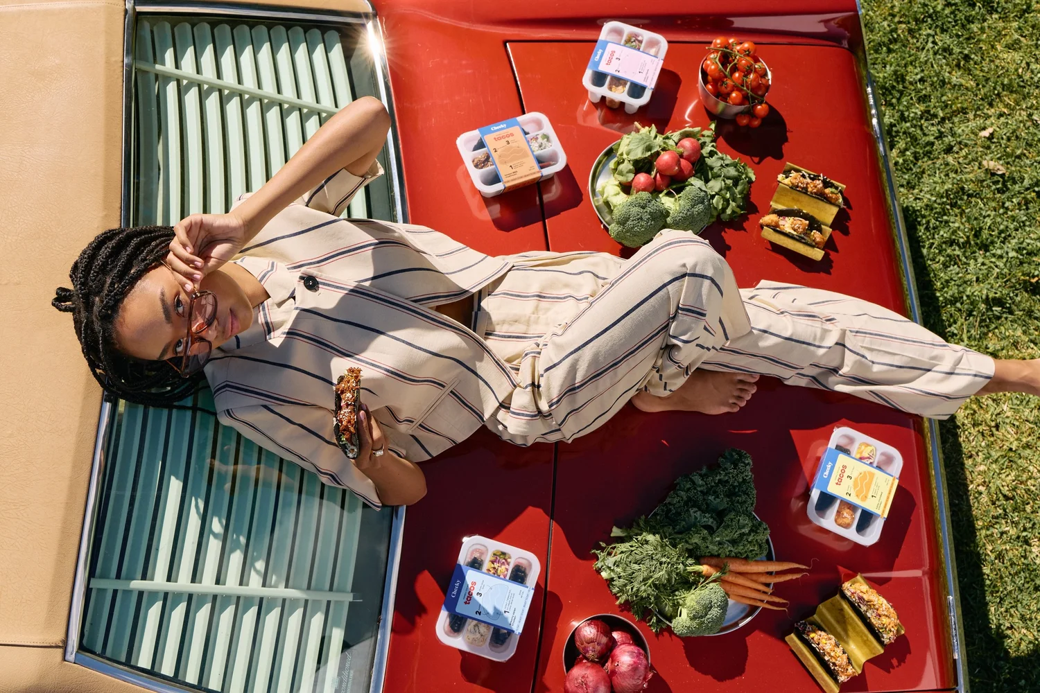 A woman enjoying a Cheeky nori taco on a red car surrounded by taco packs and fresh produce