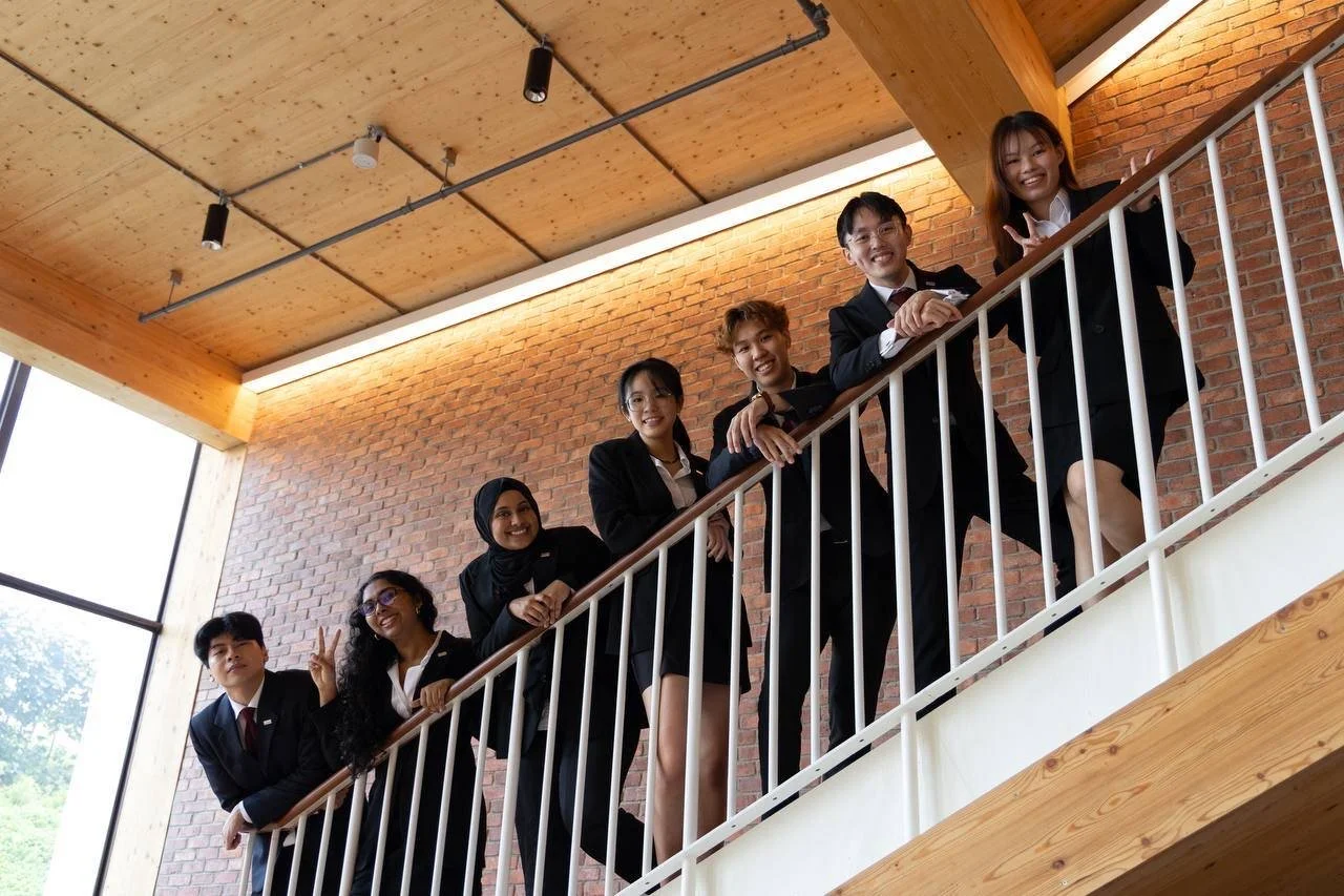 Group of seven diverse young professionals in business attire on a staircase inside a modern office building with brick wall and large windows.