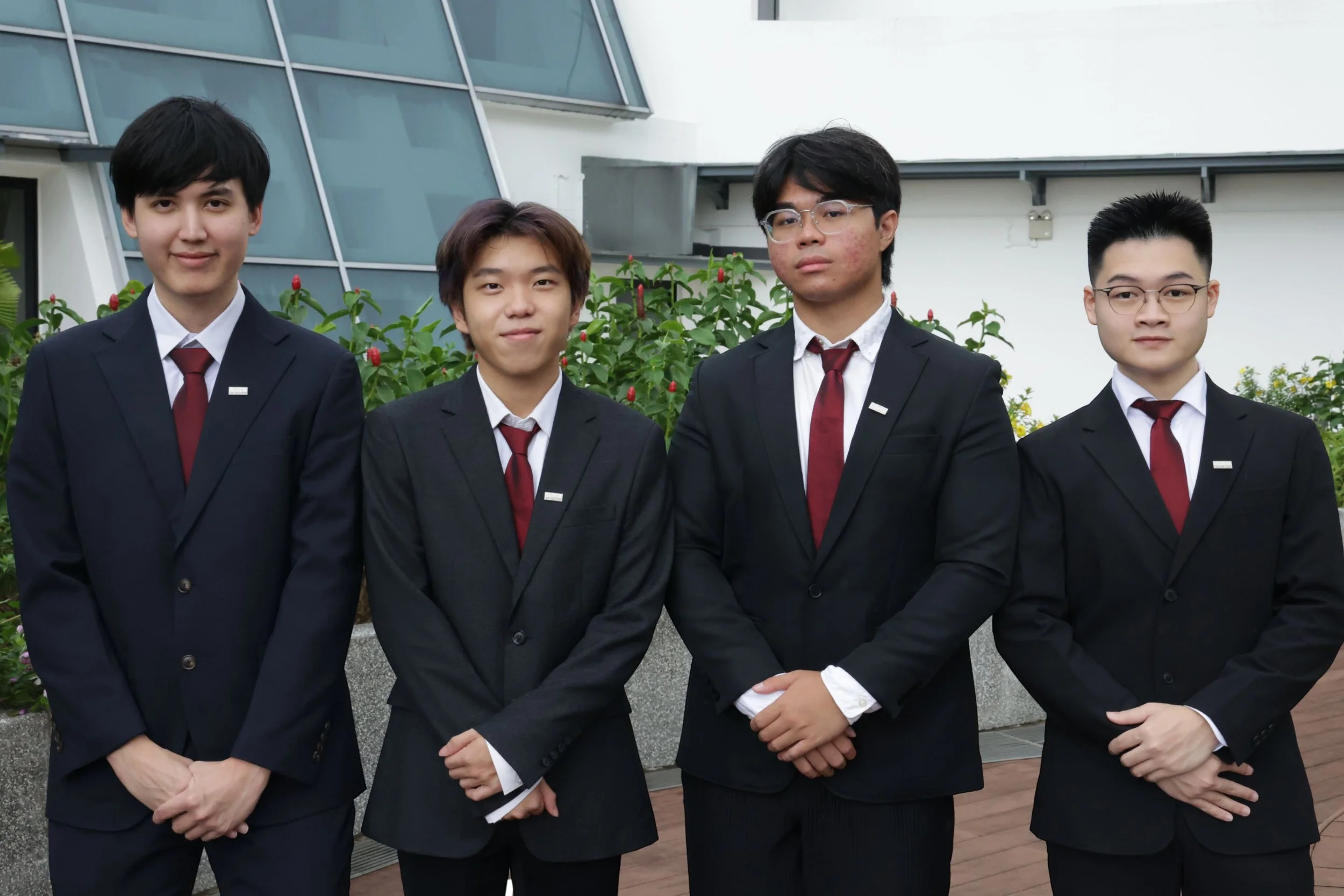 Four young men in formal black suits with white shirts and red ties standing outdoors in front of greenery and a modern building.