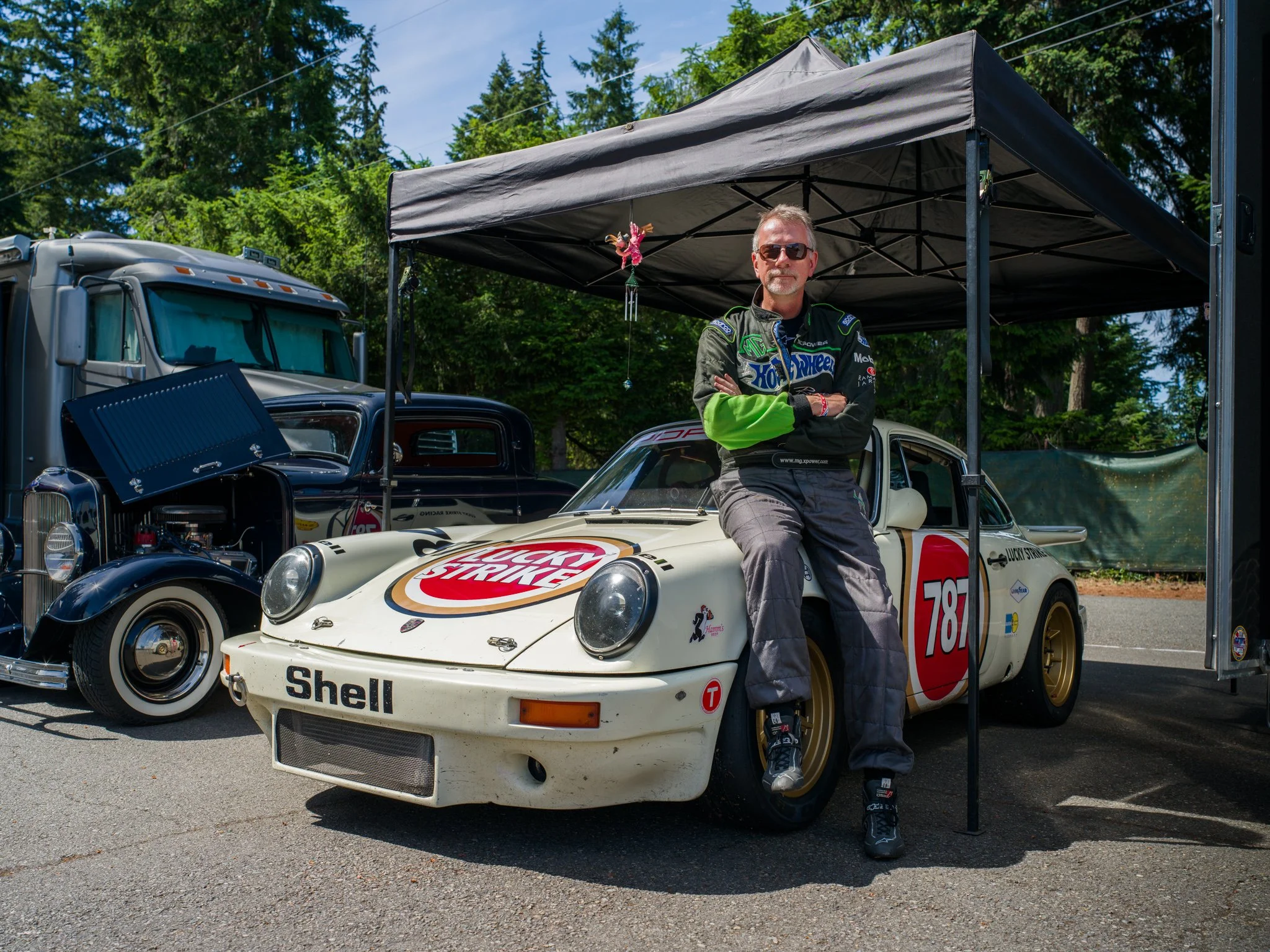 A man in racing gear sitting on a vintage race car under a canopy with trees in the background.