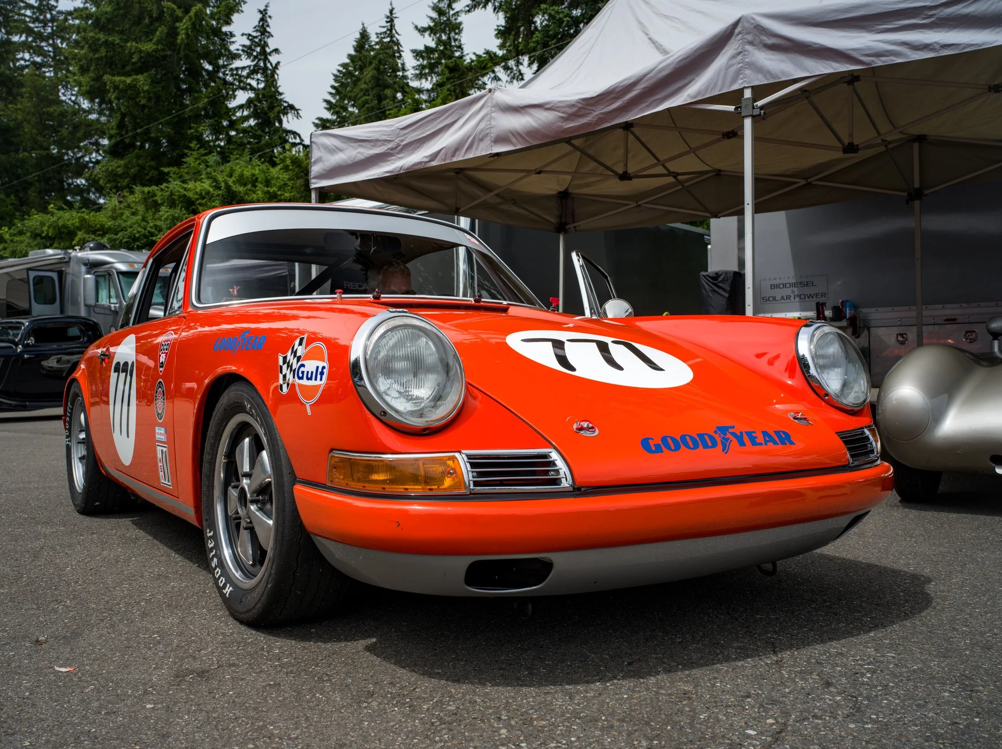 A vintage orange Porsche race car with racing decals, including Gulf and Goodyear logos, parked outdoors under a canopy with other vehicles and trees in the background.