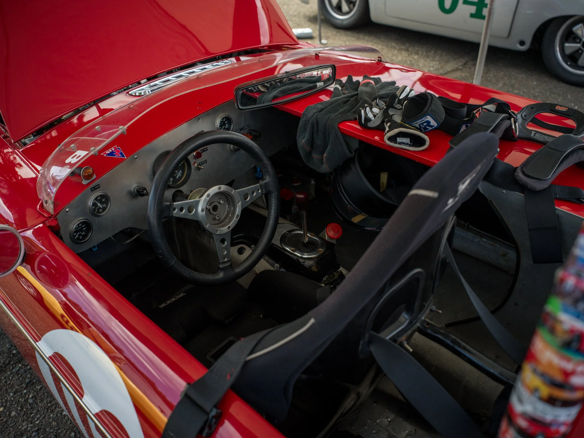 Interior of a vintage race car with a red exterior, showing a racing helmet and gloves inside, a gear shift, and racing harnesses.