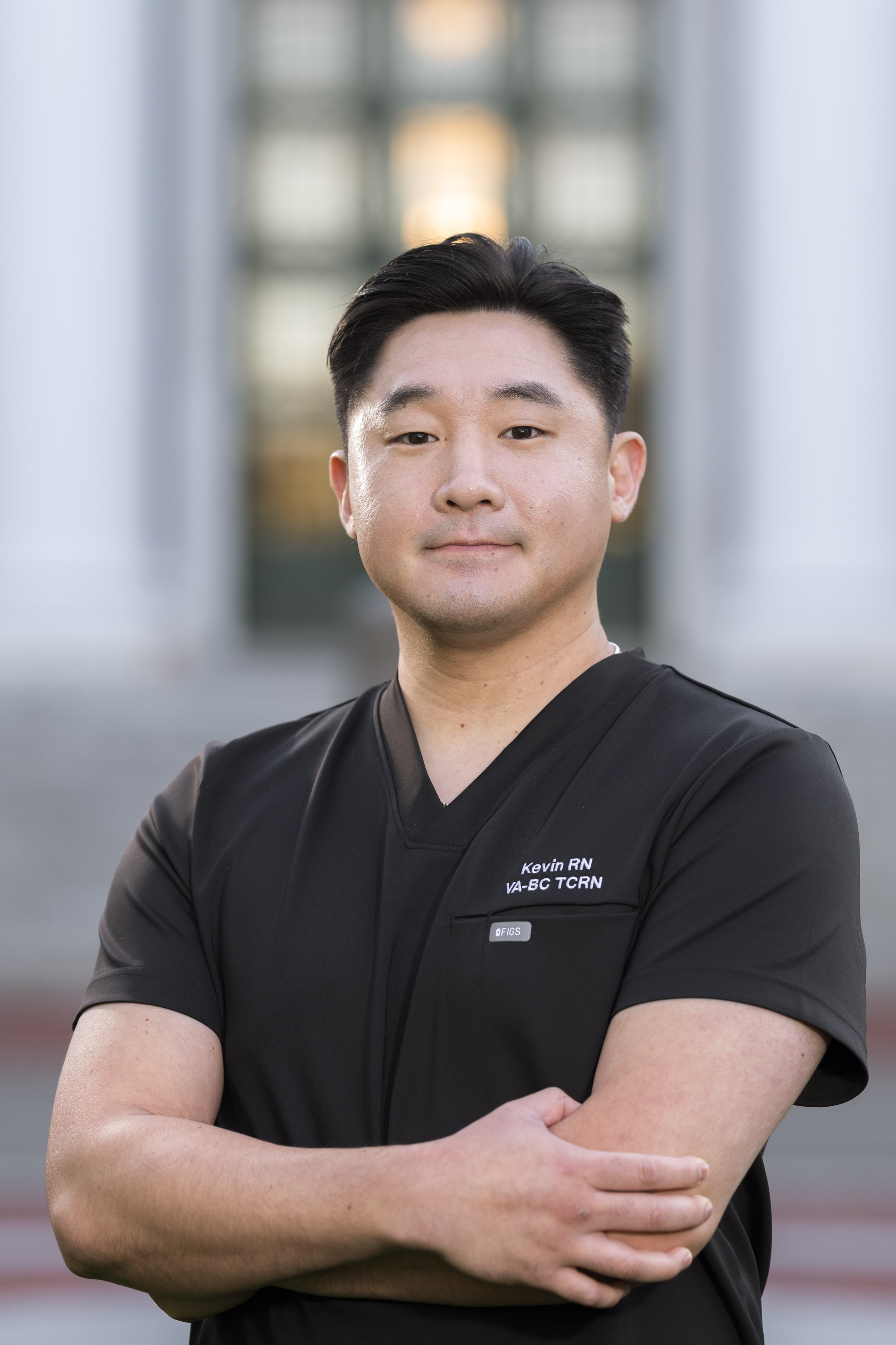 A male healthcare professional in black scrubs standing outdoors with his arms crossed, in front of a building with large windows.