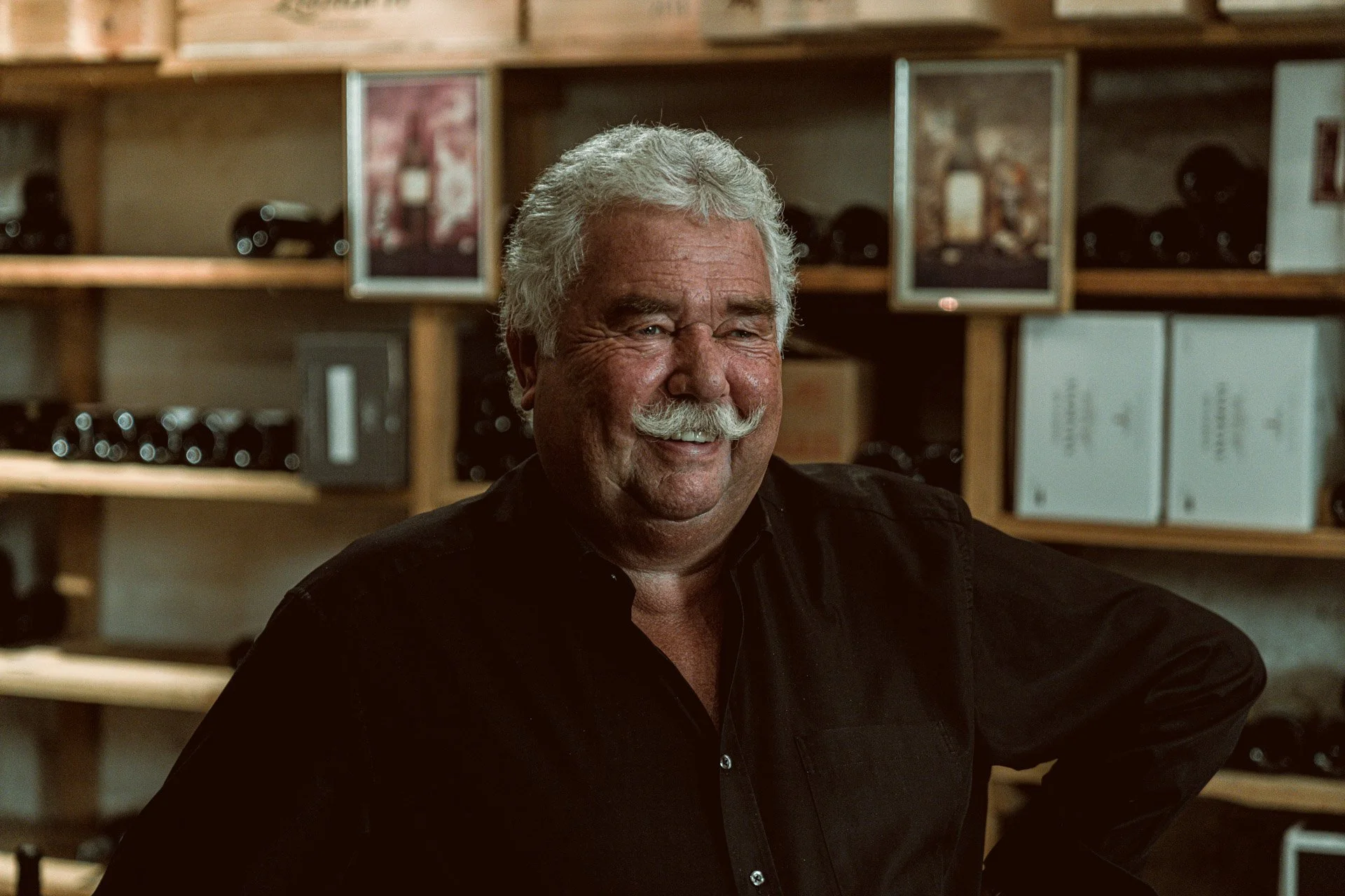 Älterer Mann mit Schnurrbart steht vor einem Weinregal.