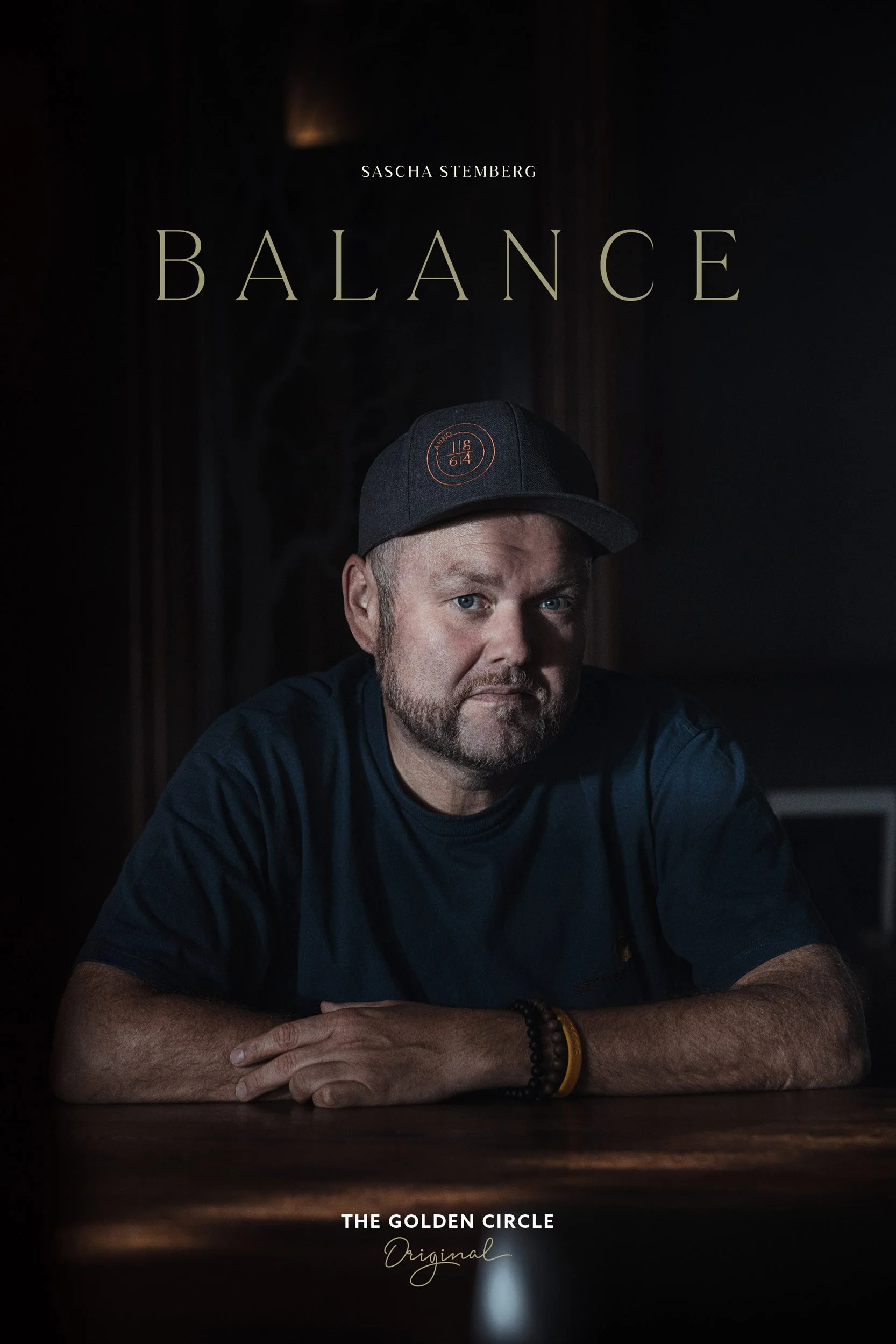 Das Bild zeigt das Cover eines Buches oder Films mit dem Titel 'Balance' von Sascha Stemberg. Ein Mann mit einer Kappe sitzt an einem Tisch und schaut in die Kamera. Unten steht 'The Golden Circle Original'. Die Szene hat eine dunkle, atmosphärische Beleuchtung.