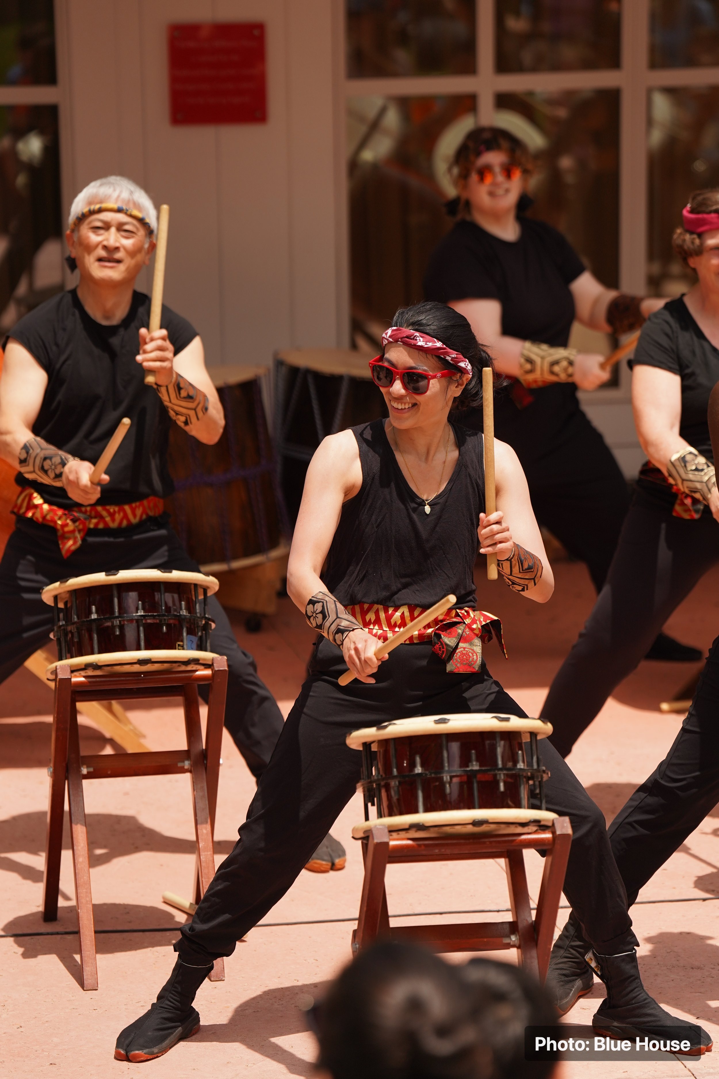 Group of four people performing a Japanese taiko drumming routine, wearing black outfits, headbands, and sunglasses outdoors.
