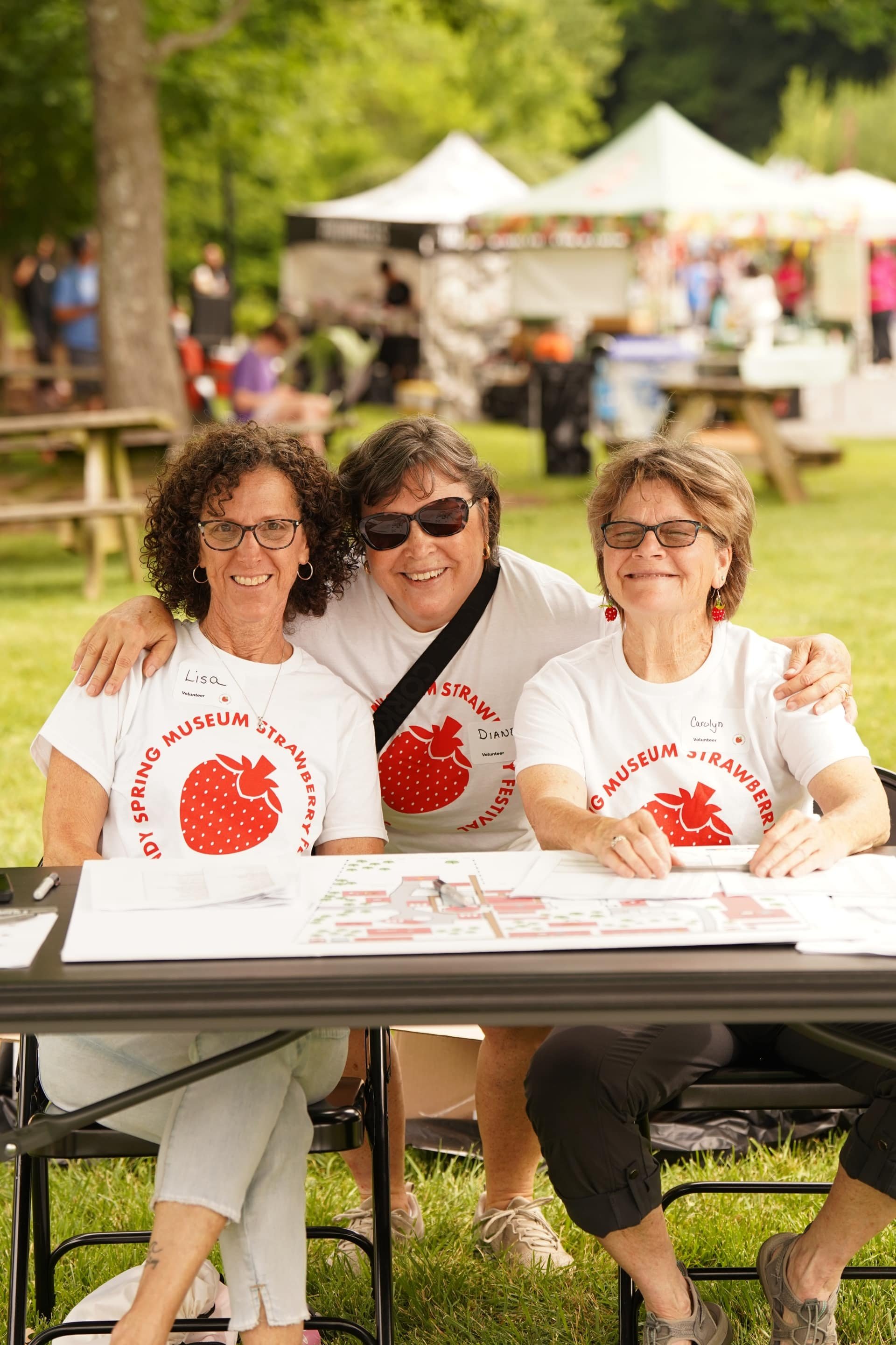 Three volunteers smiling at a welcome table during a strawberry-themed outdoor festival.