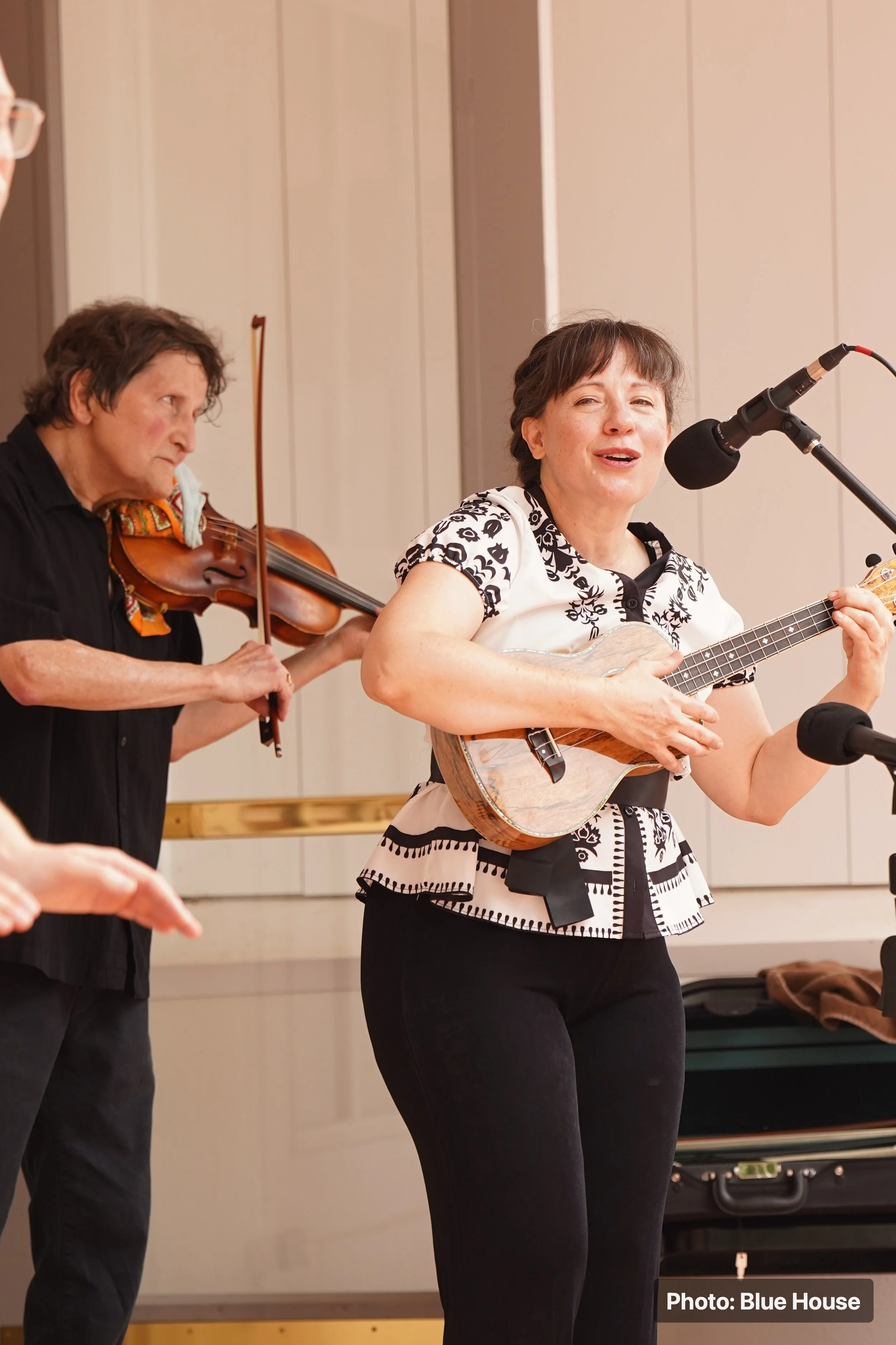 Two women performing music; one playing a violin, the other strumming a guitar and singing into a microphone.