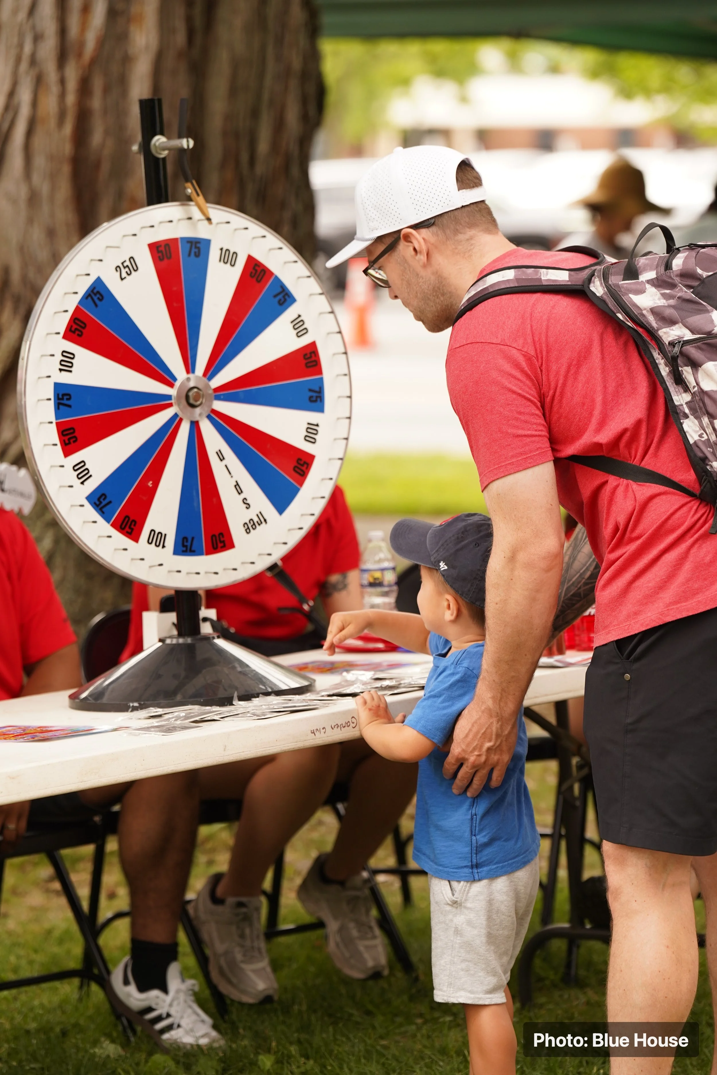 A man and a young boy playing a game of Lucky Ducks at an outdoor event. The game features a spinning wheel with red and blue sections, and there are people sitting at a table behind them.