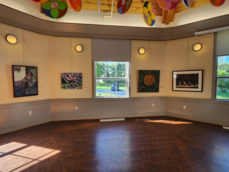 Interior of an art gallery with five paintings on the walls. The room has wooden flooring and colorful decorations hanging from the ceiling. Sunlight streams through large windows.