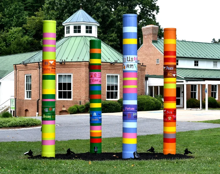 Colorful cylindrical sculptures with positive words like "Hope," "Love," and "Peace" in English and Chinese characters stand on a grassy area in front of a brick building with a green roof.