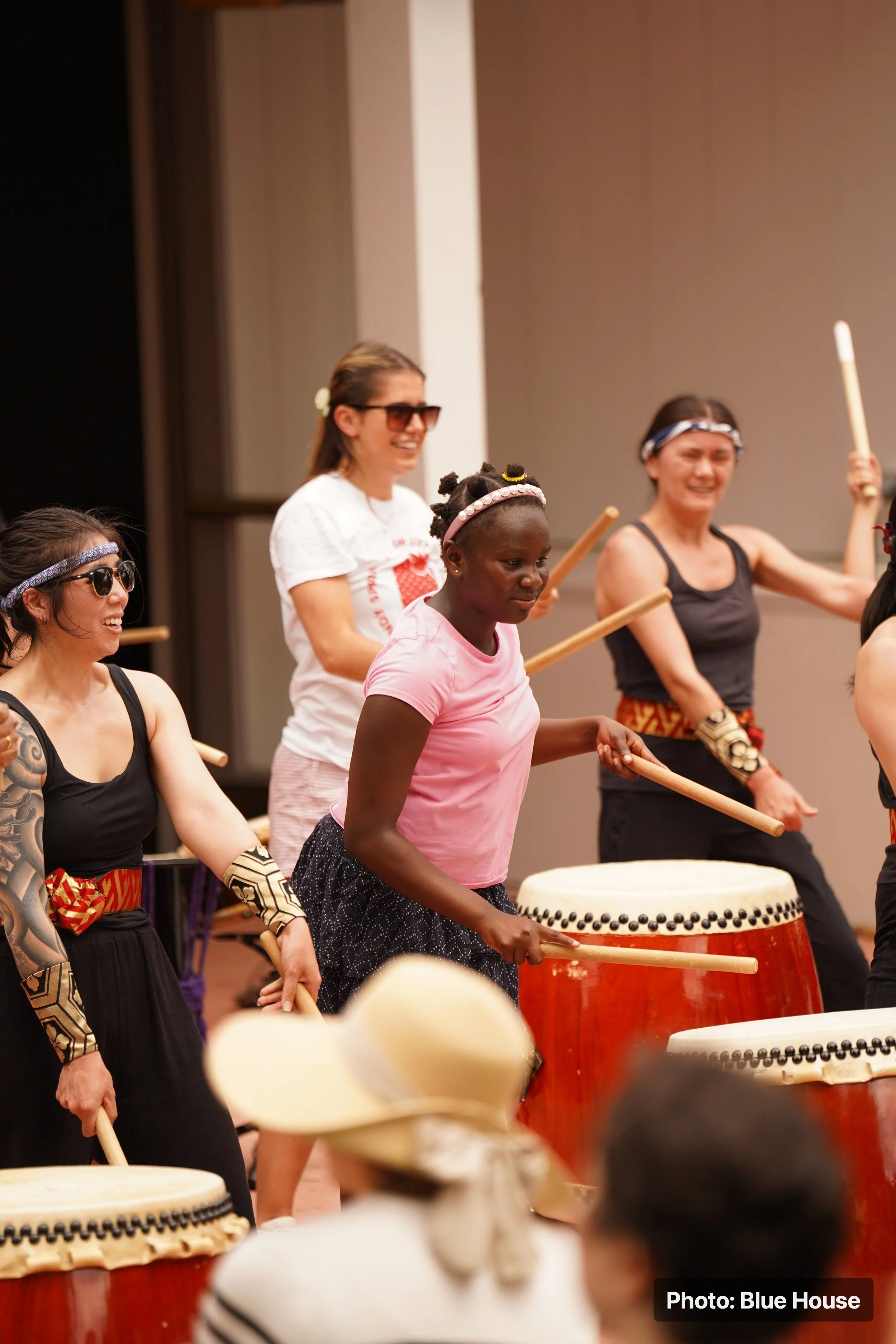 Group of women and girls playing traditional drums with sticks during the festival, dressed in casual and traditional attire.