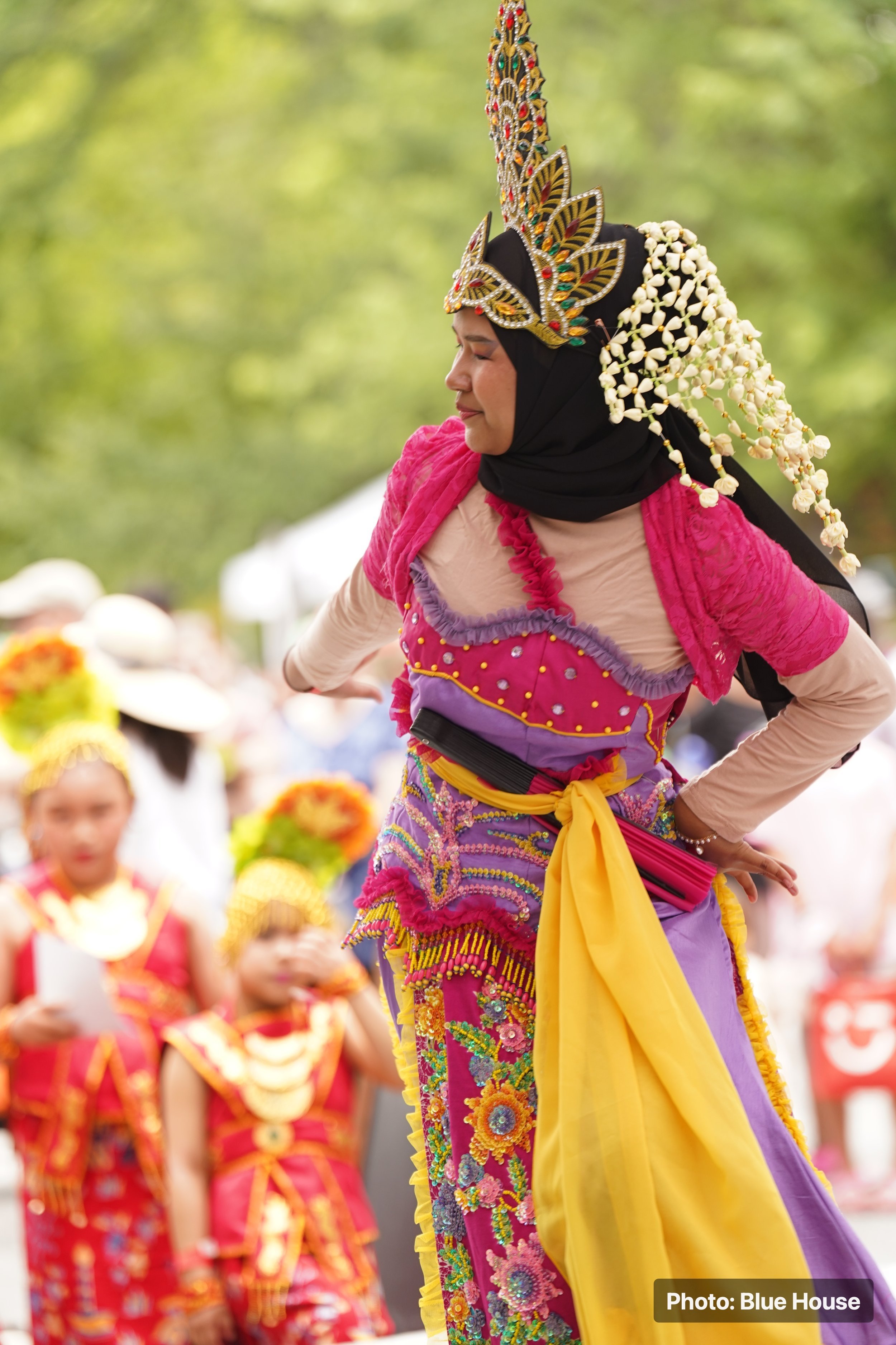 A woman dressed in traditional, colorful attire is dancing during the festival, with a group of similarly dressed women in the background.