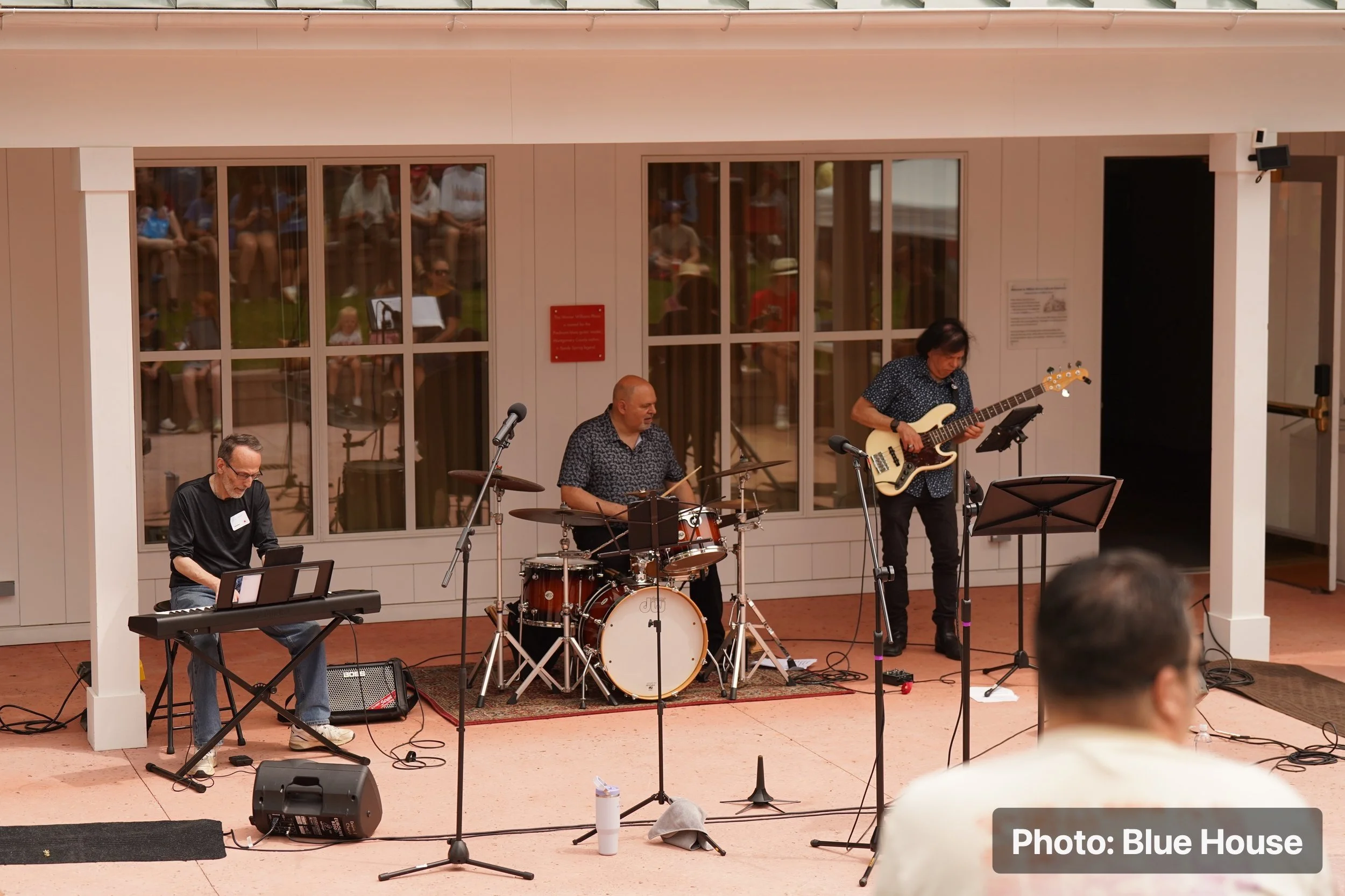 A band performing live music outdoors, featuring a keyboard player, a drummer, and a bassist, with audience members visible through large glass windows behind them.