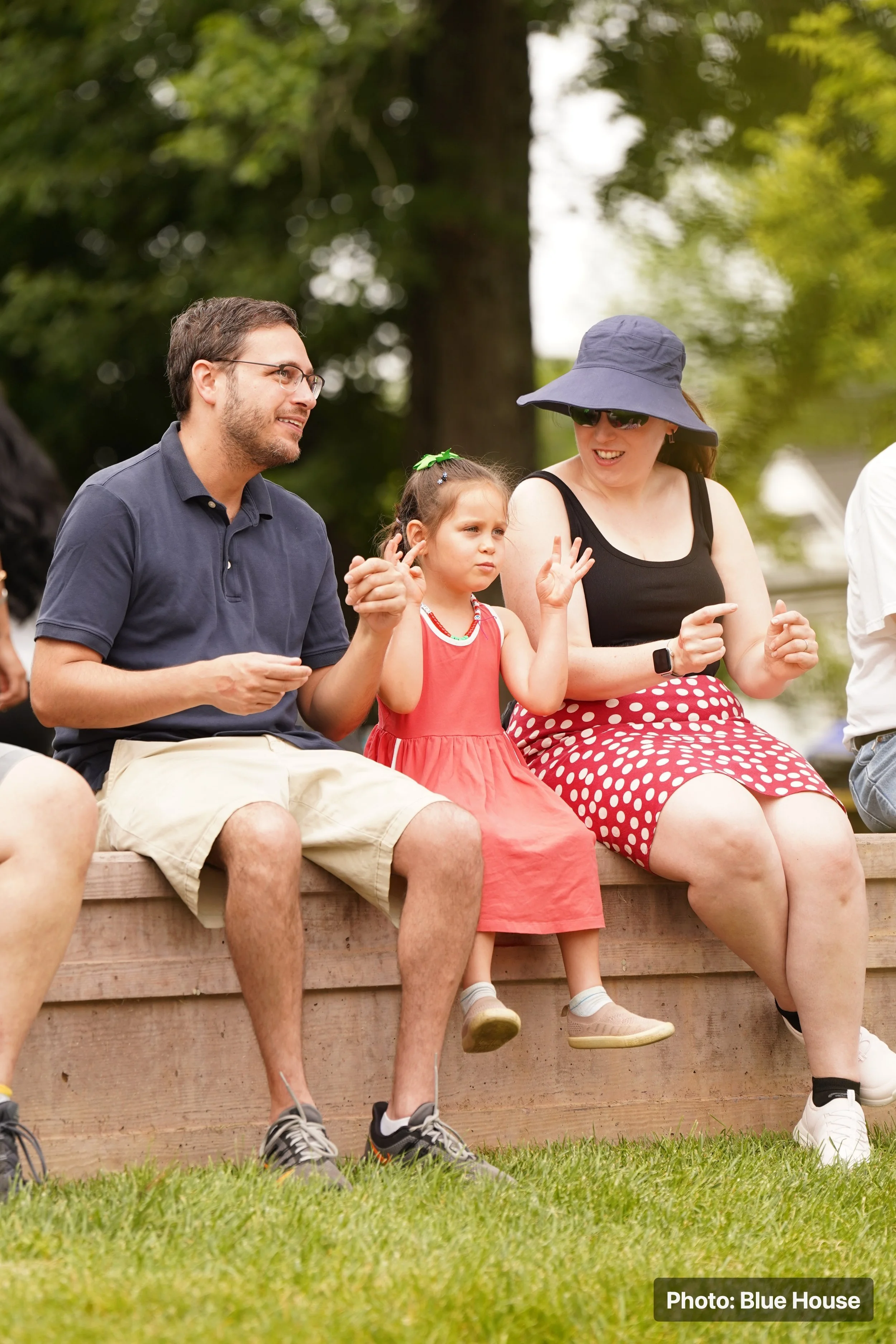 A family sitting outdoors on a wooden bench in a park with green trees in the background. There is a man with glasses and a navy shirt, a young girl in a red dress, and a woman wearing a wide-brimmed hat, sunglasses, and a polka-dot skirt, all engage