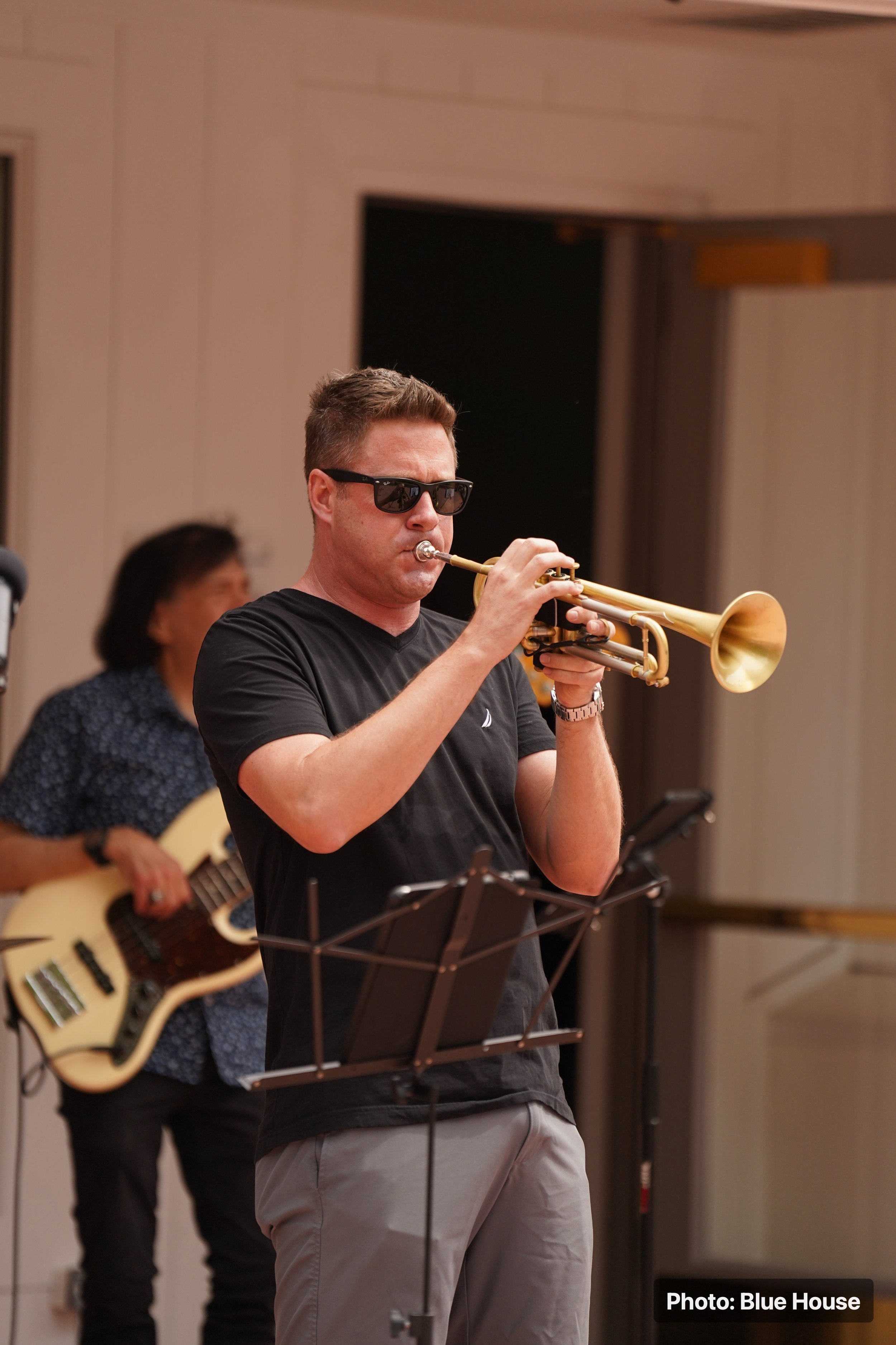 A man wearing sunglasses and a black t-shirt playing a trumpet, with a woman in the background playing an acoustic guitar in an indoor setting.