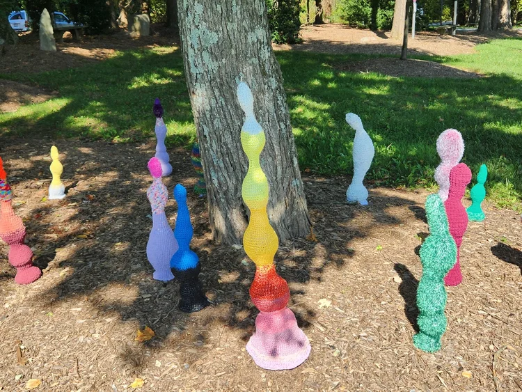 Colorful, abstract crochet sculptures arranged on the ground beneath a tree in an outdoor park setting.