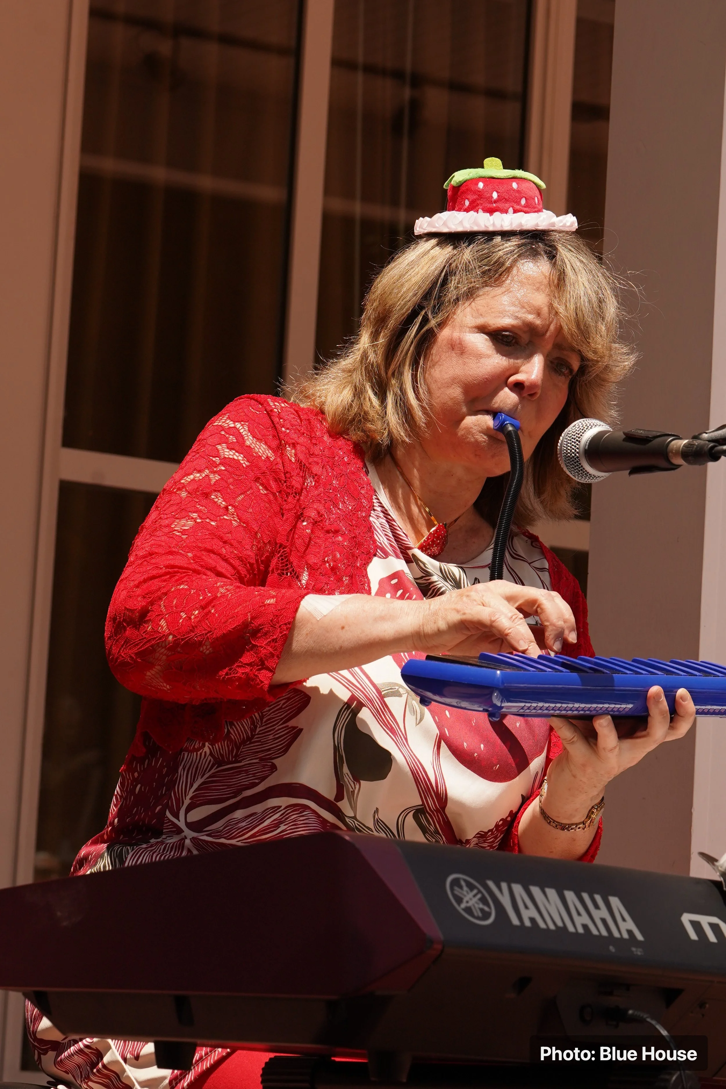 A woman with blonde hair is playing a Yamaha keyboard. She is wearing a dress with a strawberry pattern and a red lace jacket. She has a strawberry-shaped hat on her head.