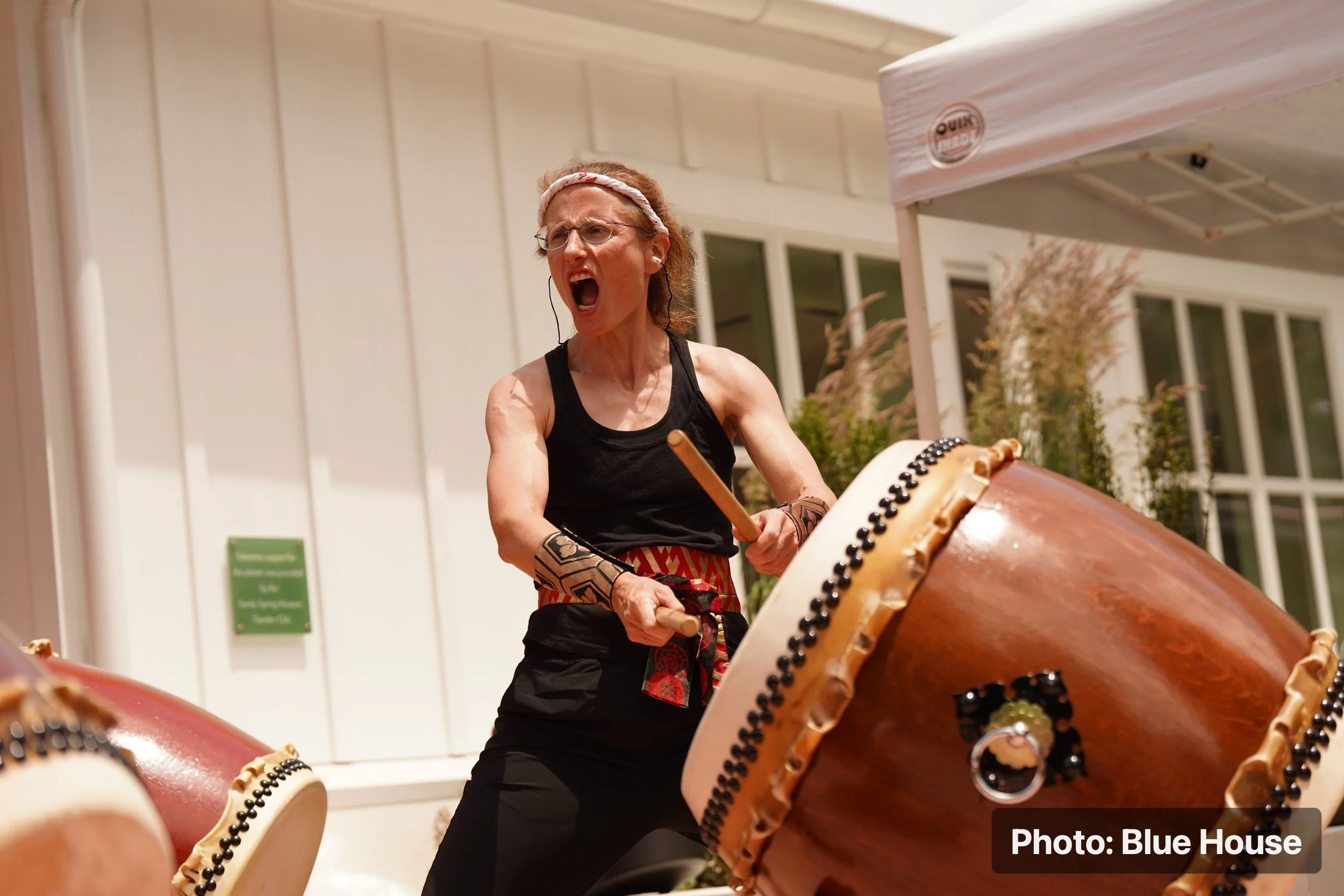 An older woman with glasses, tattoos, and earrings passionately plays a large traditional drum outdoors. She is wearing a black sleeveless top, a patterned headband, and a red patterned sash, with her mouth open in an expressive act of music.