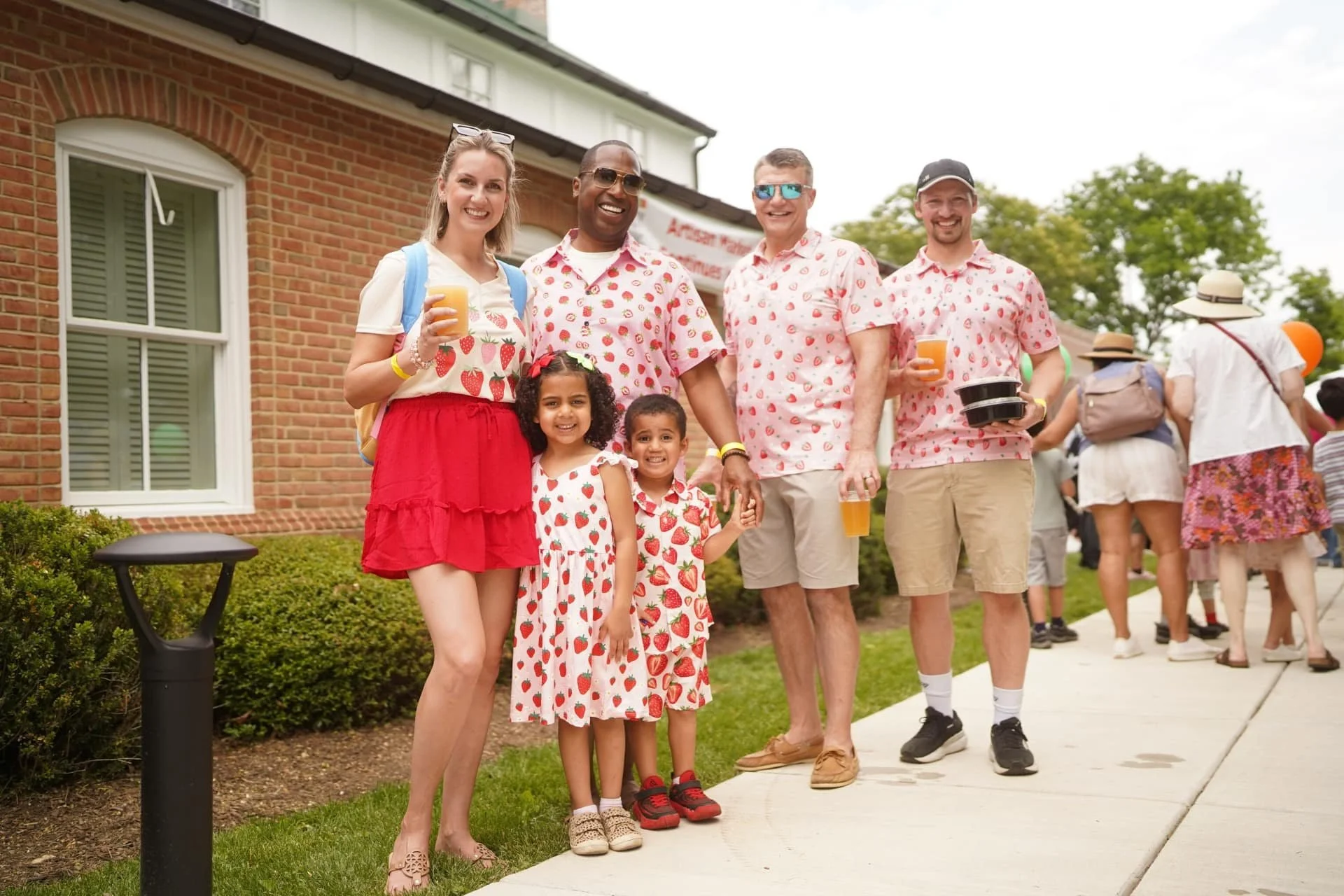 A group of families and visitors posing in strawberry-themed outfits outside.