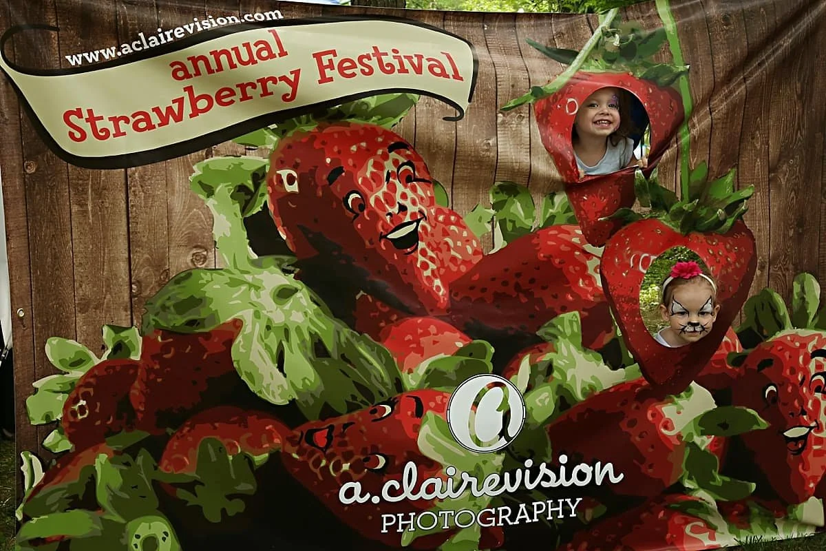 Visitors posing in a strawberry-themed photo installation at the Strawberry Festival, featuring large illustrated strawberries and playful cutouts.