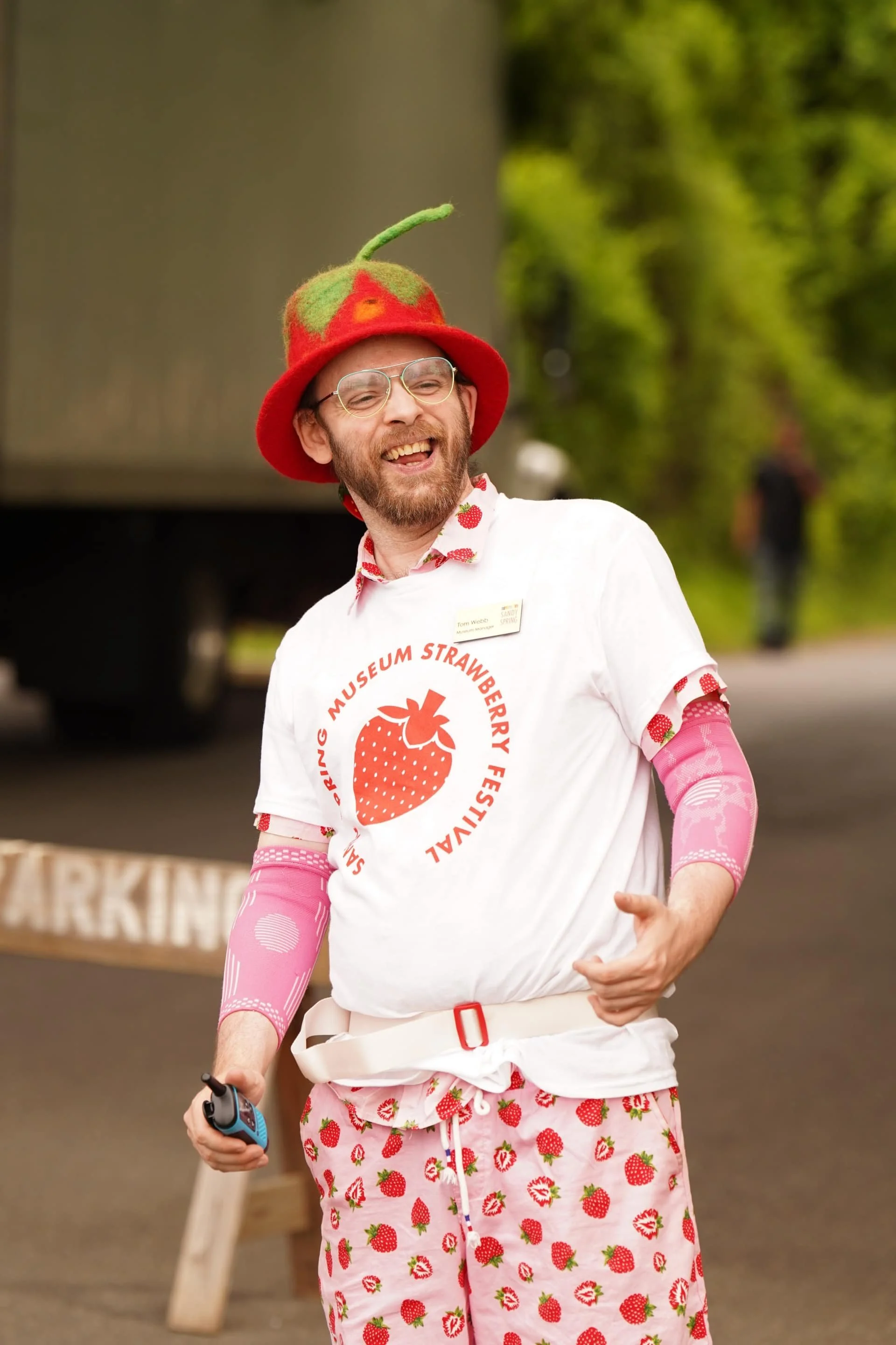 A museum staff wearing strawberry-themed clothing and a strawberry hat smiles outdoors.