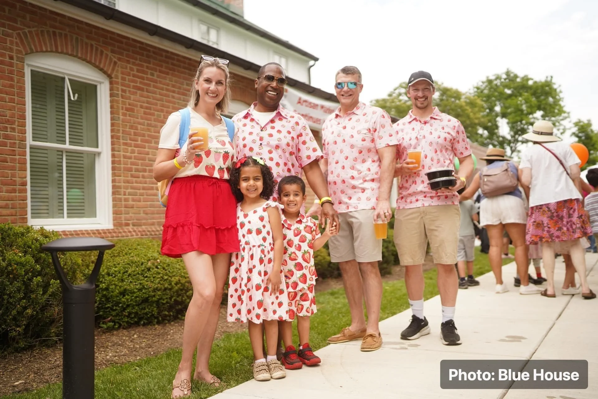 A group of families and visitors posing in strawberry-themed outfits outside.