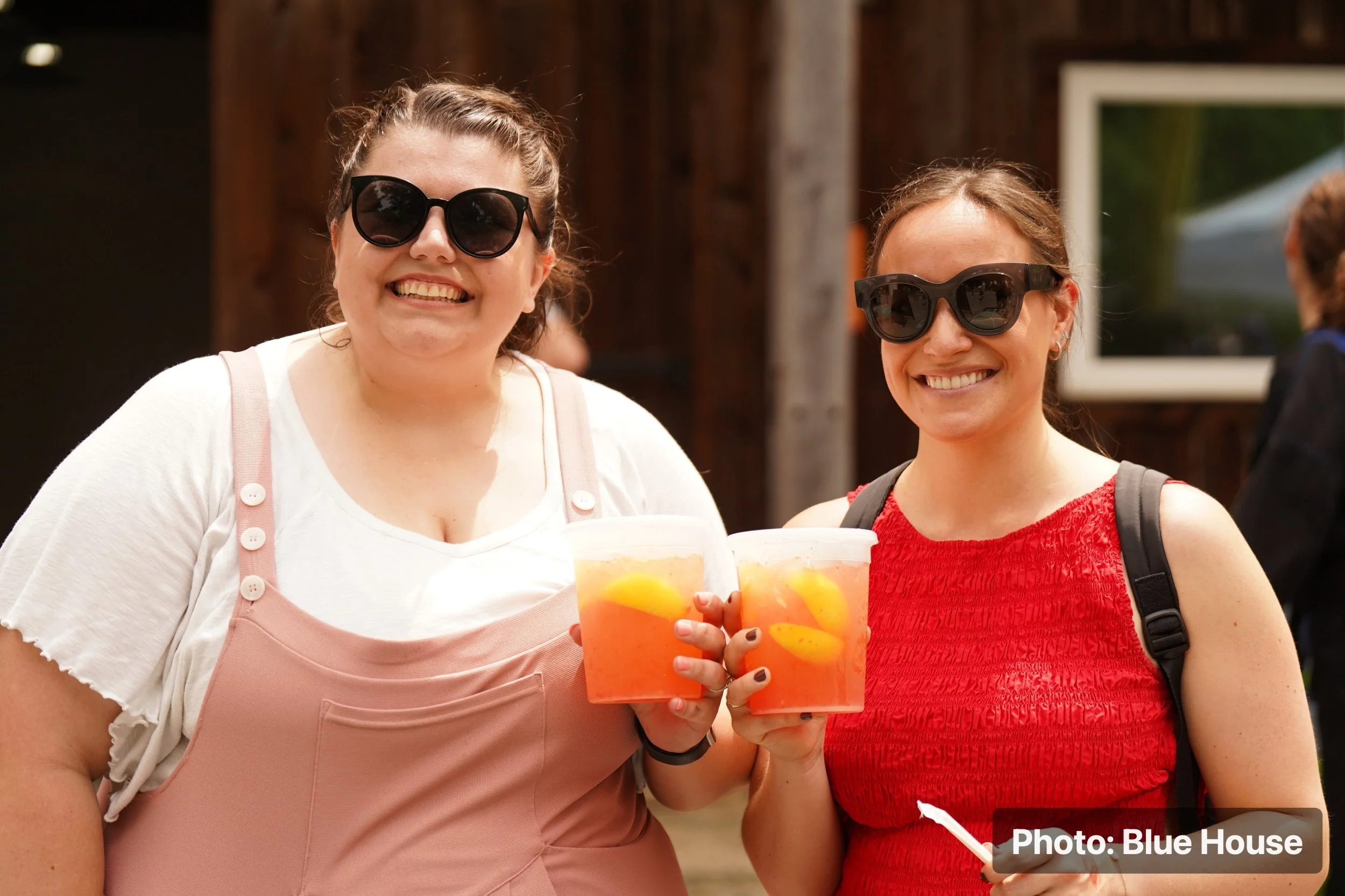 Two smiling women wearing sunglasses, holding drinks, and standing outdoors.
