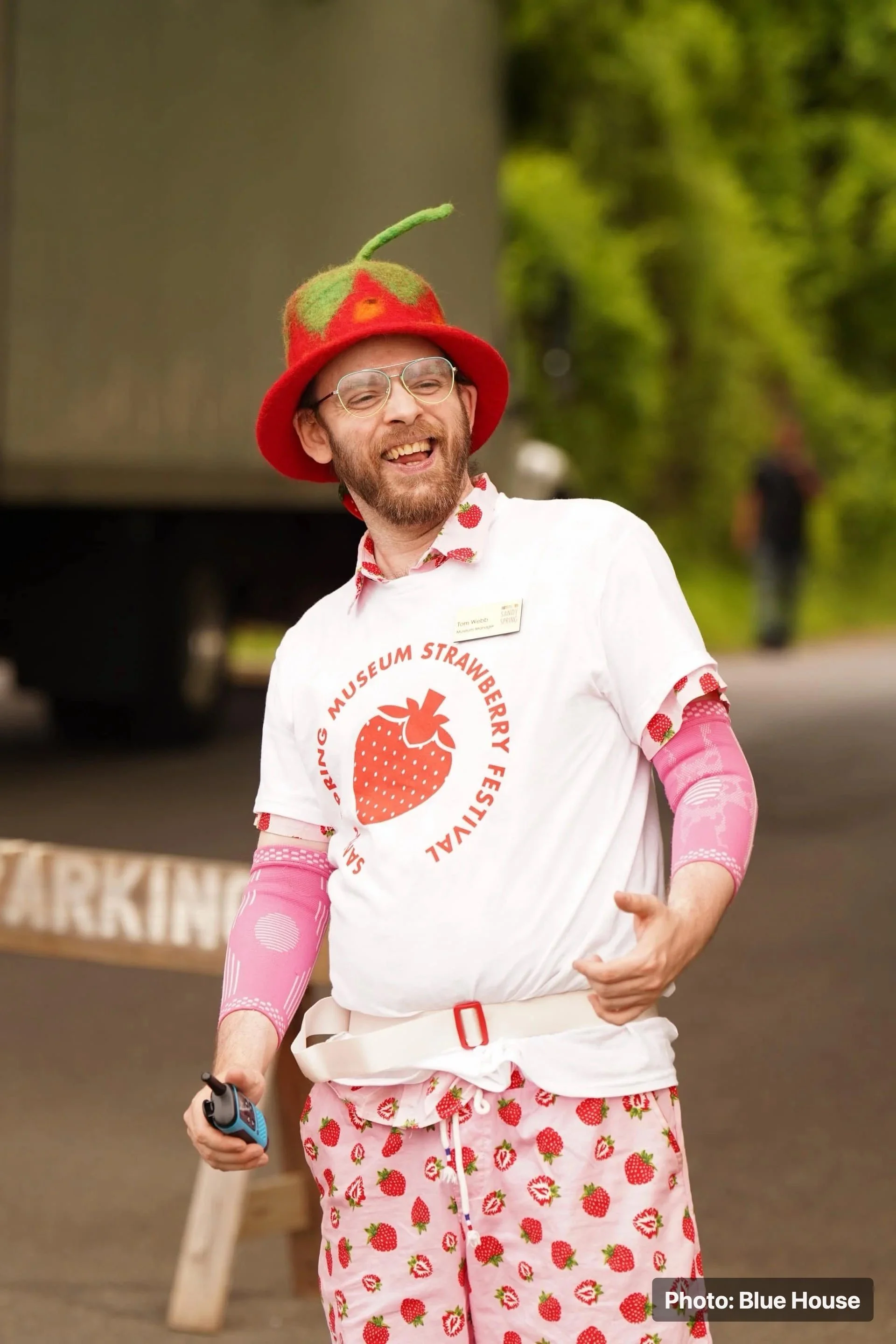 A museum staff wearing strawberry-themed clothing and a strawberry hat smiles outdoors.