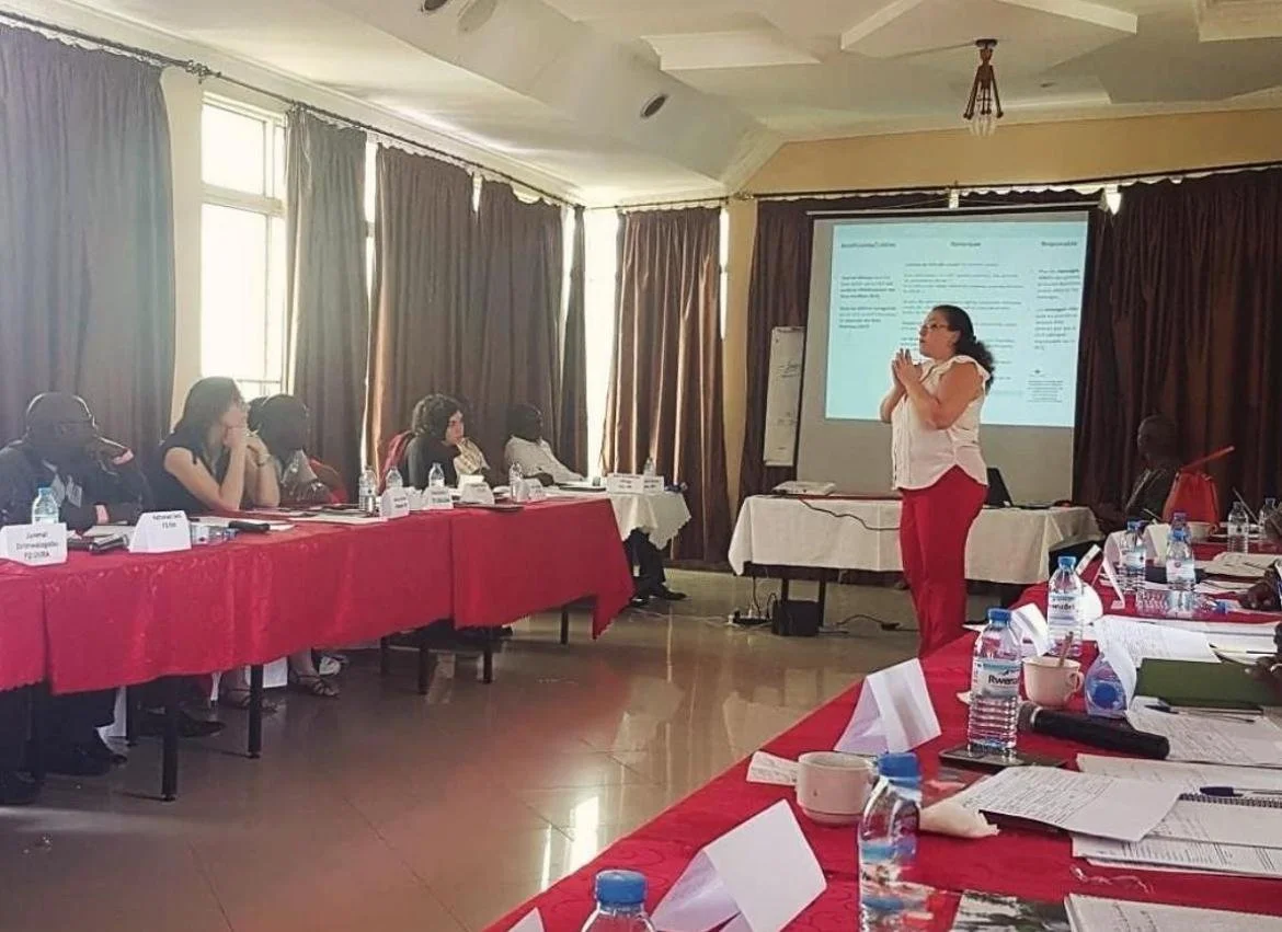 Chahrazed Anane, founder of Humanitarian Career Consulting giving a presentation to a group of people seated at a conference table in a room with large windows and brown curtains in Kinshasa