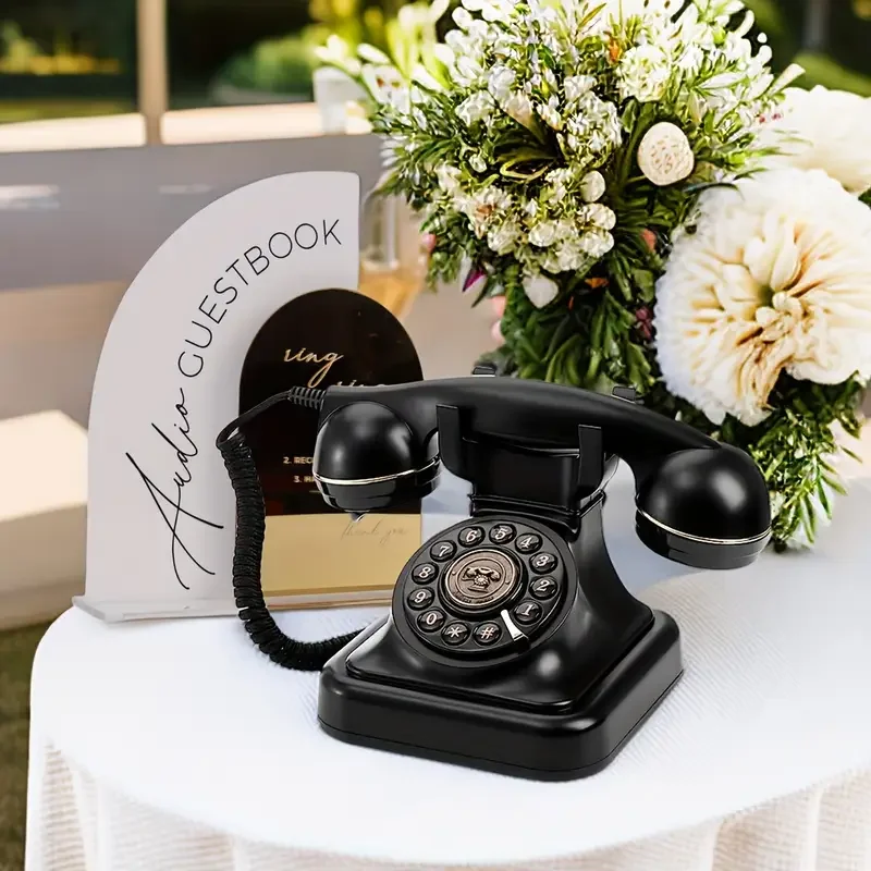 Vintage black rotary dial telephone on a white table, with a floral arrangement and a guestbook in the background.