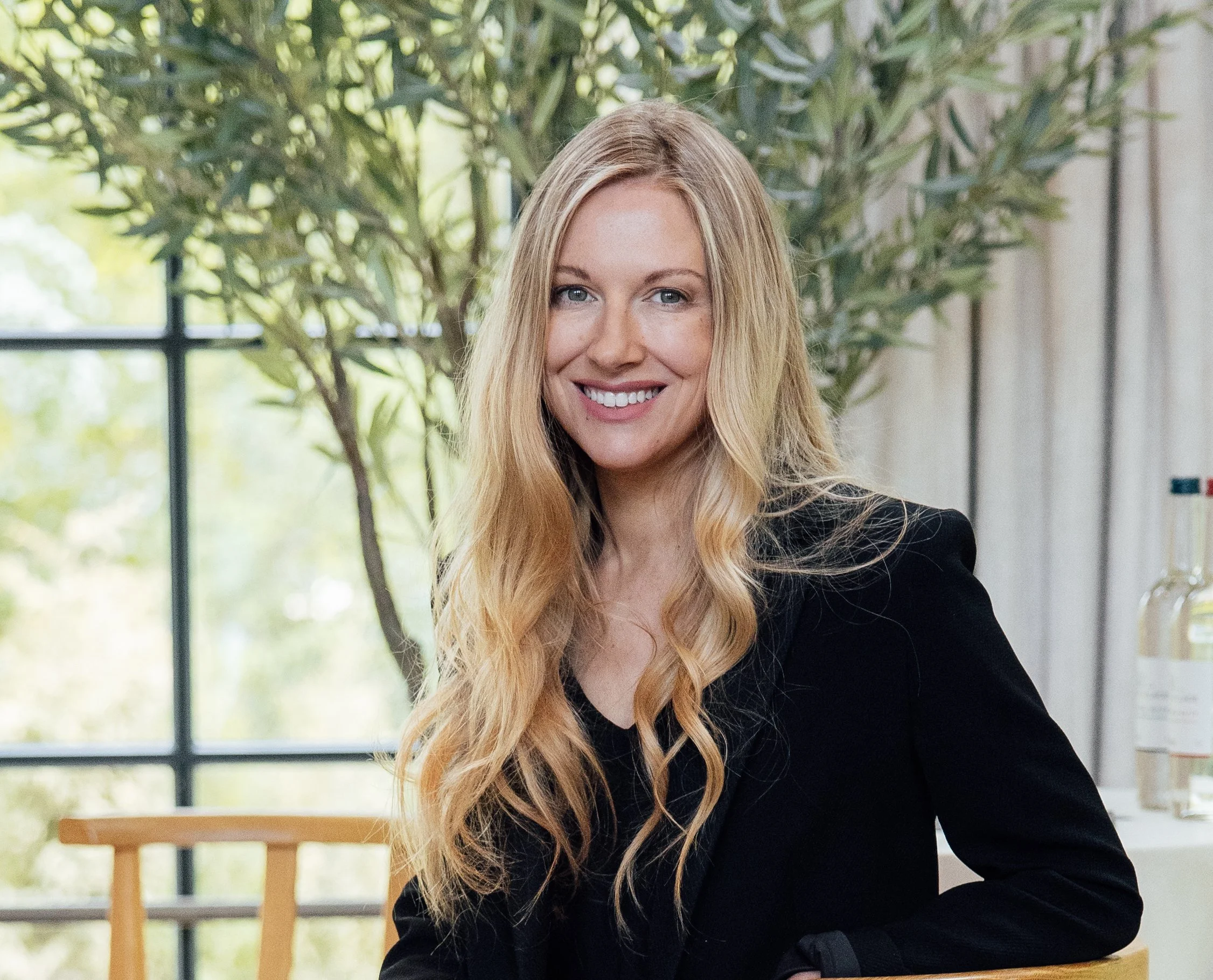 A woman with long blonde hair and a black blazer sitting indoors near a window with plants in the background, smiling at the camera.