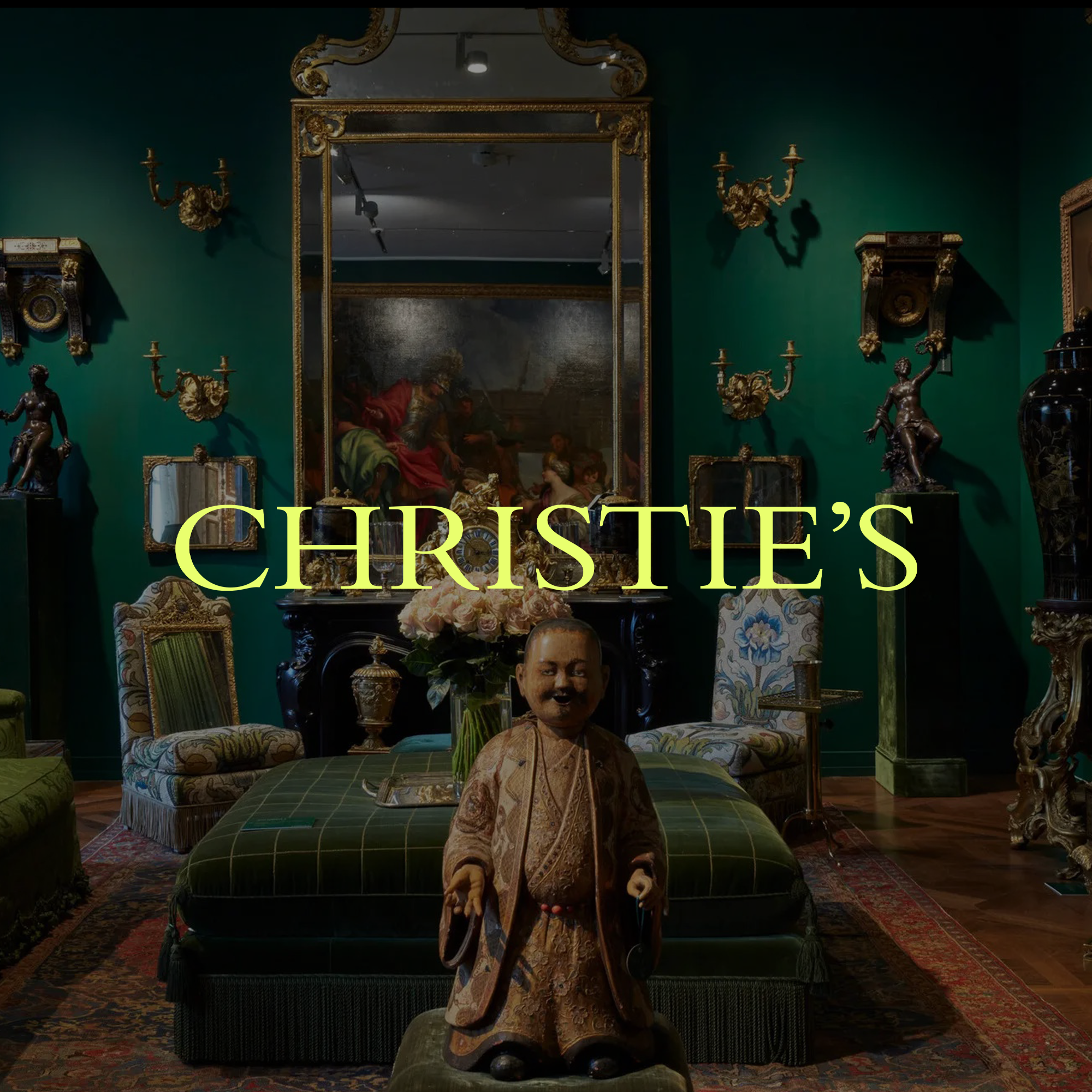 An art gallery room with green walls, antique furniture, and ornate gold-framed mirrors and sculptures. In the center, a wooden figure with a painted face and traditional clothing stands in front of a floral centerpiece on a green-tufted ottoman. The room features various decorative items and a large mirror reflecting a classical painting.