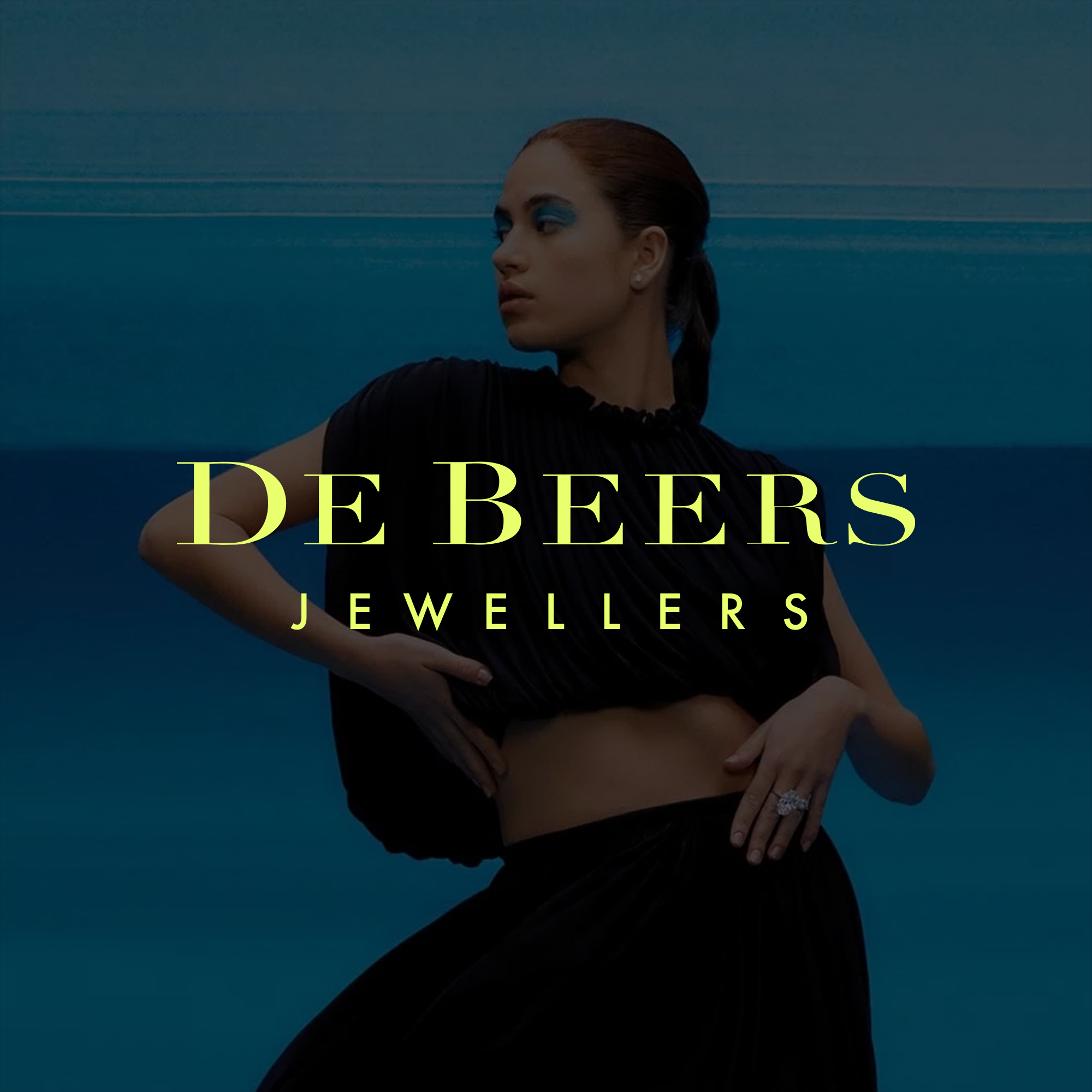 A woman with dark hair in a low ponytail wearing a black outfit with a ring on her finger, standing against a blue background with horizontal lines, overlaid with the text "DE BEERS JEWELLERS."