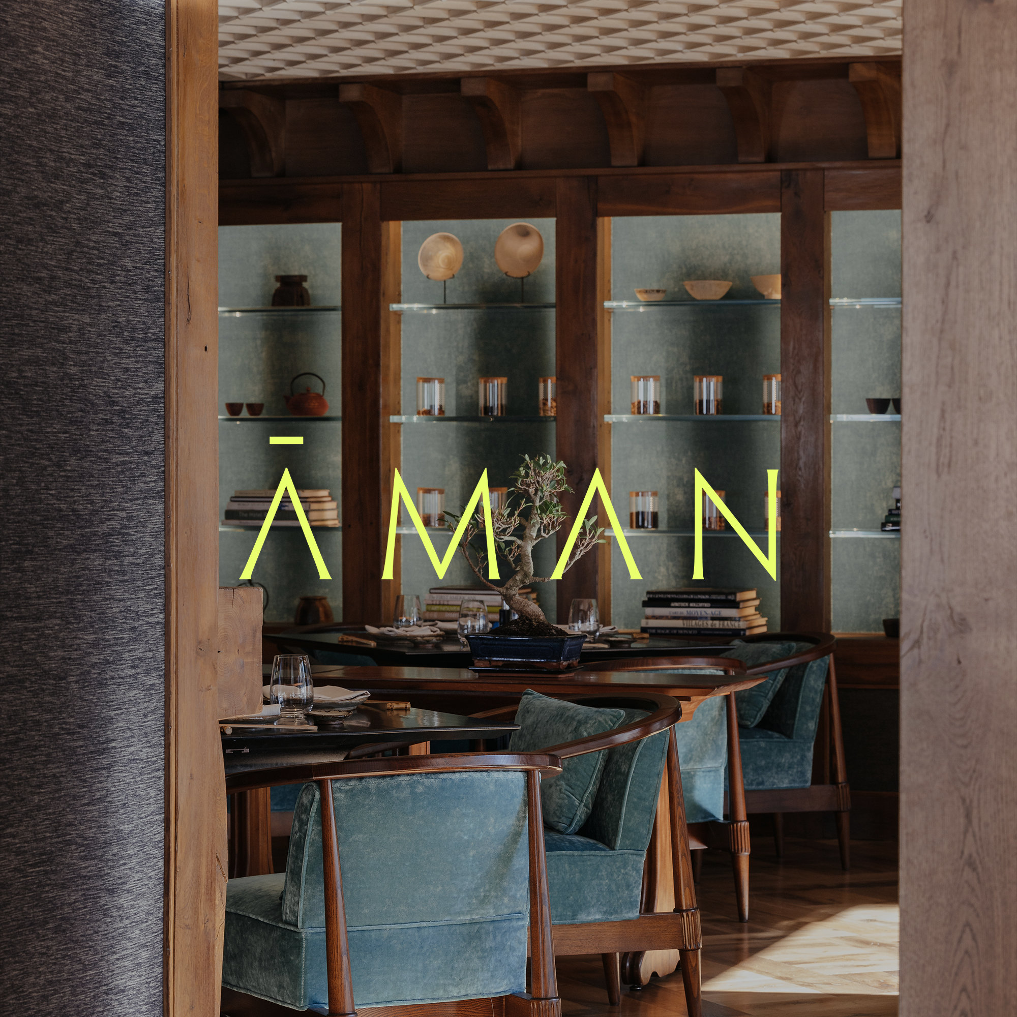 View of a restaurant interior with green upholstered chairs, a wooden table with a bonsai tree centerpiece, and wooden shelving with pottery and books. The word 'ÁMAN' is written across the window in yellow neon letters.