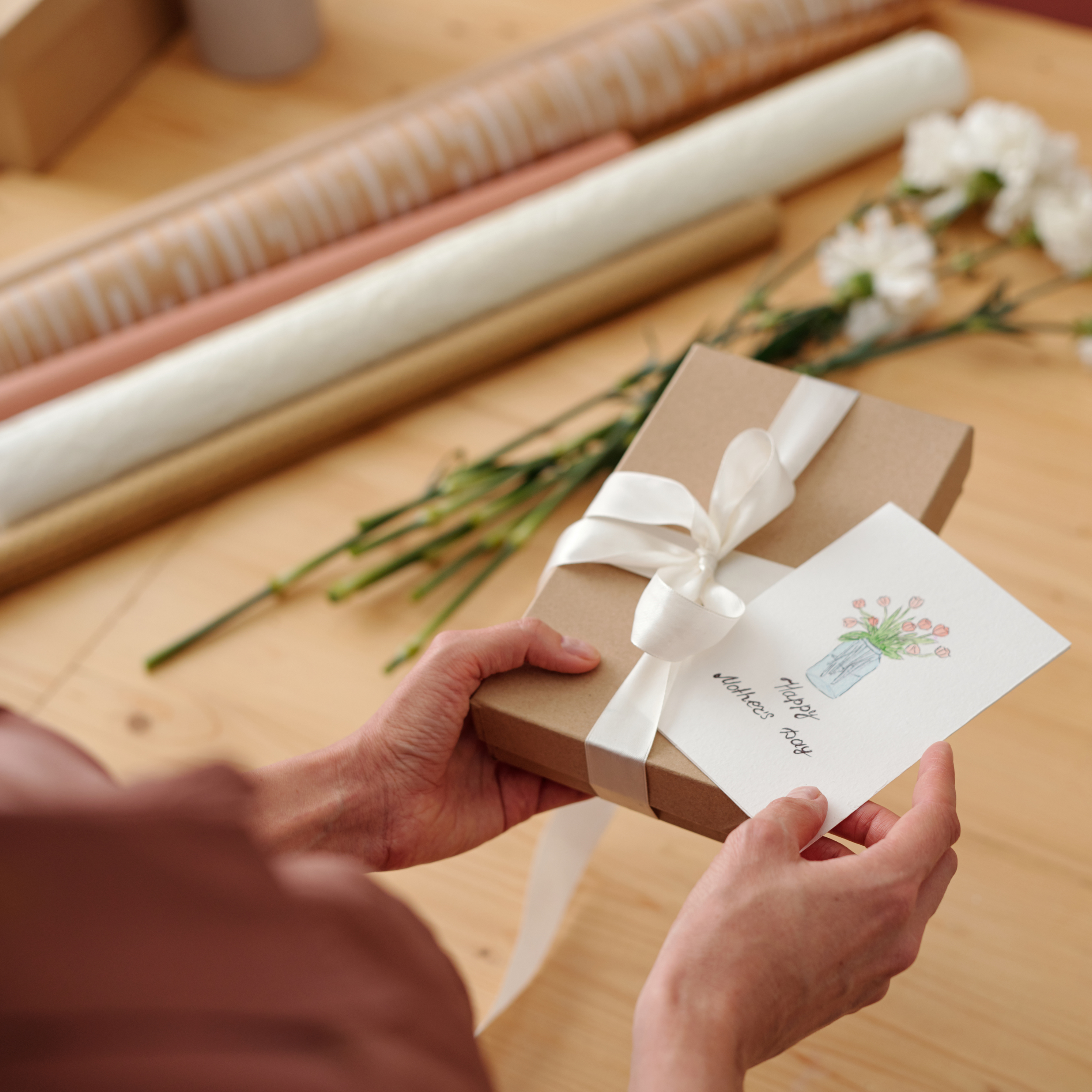 A person holding a wrapped gift box with a white ribbon, a greeting card with a flower drawing, and white flowers on a wooden table, with wrapping paper rolls in the background.