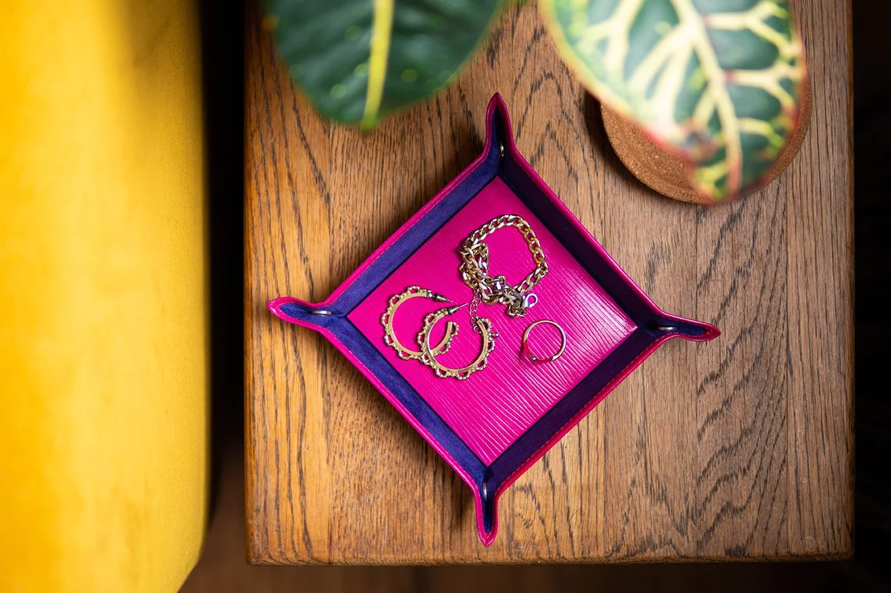 A fuchsia jewellery tray with gold jewellery, including a bracelet, two hoop earrings, and a ring, sitting on a wooden surface next to a green potted plant.