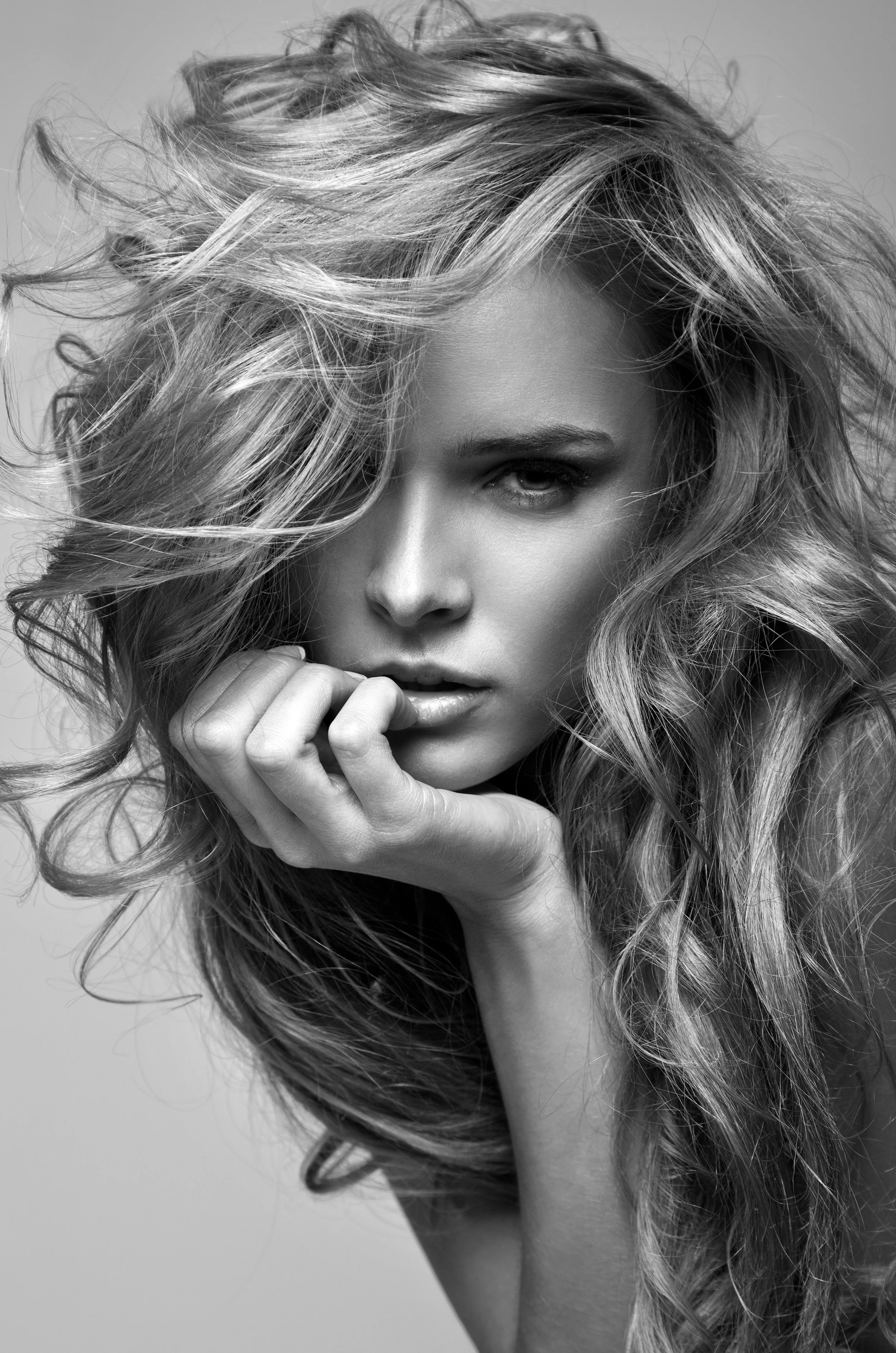 Photographie en noir et blanc d'une femme aux cheveux ébouriffés regardant l'appareil photo, le visage partiellement couvert et la main sous le menton.