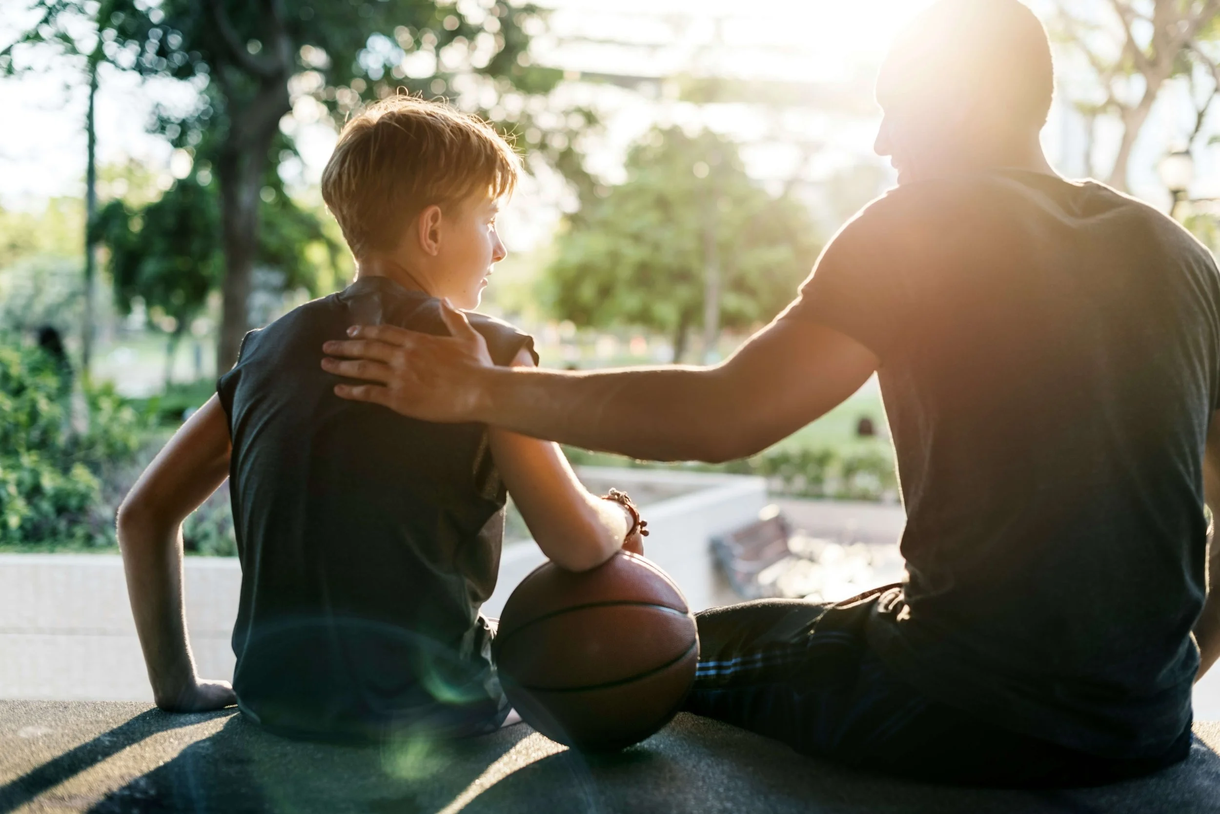 Father and son sitting together outdoors with a basketball in warm sunlight