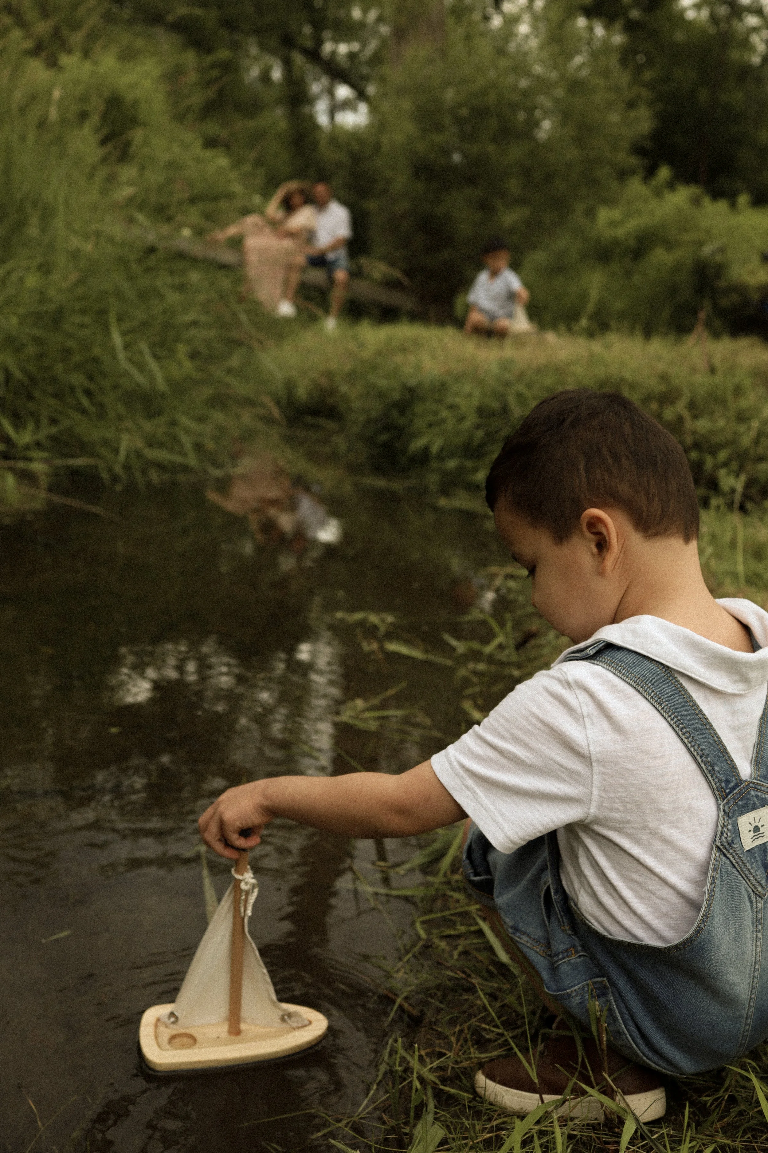Young boy crouched by a creek playing with a toy sailboat, with three people sitting on a bench in the background in a green outdoor setting.