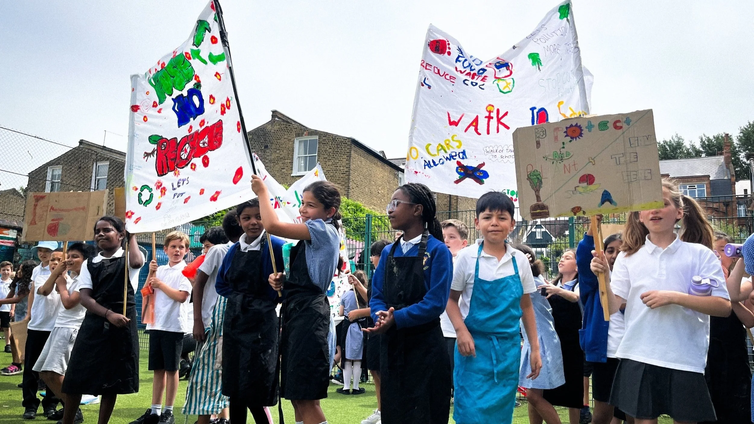 Retrofit Action For Tomorrow — Primary school students stand outside on artificial grass, holding up banners and signs that they have made sharing their ambitions for a sustainable