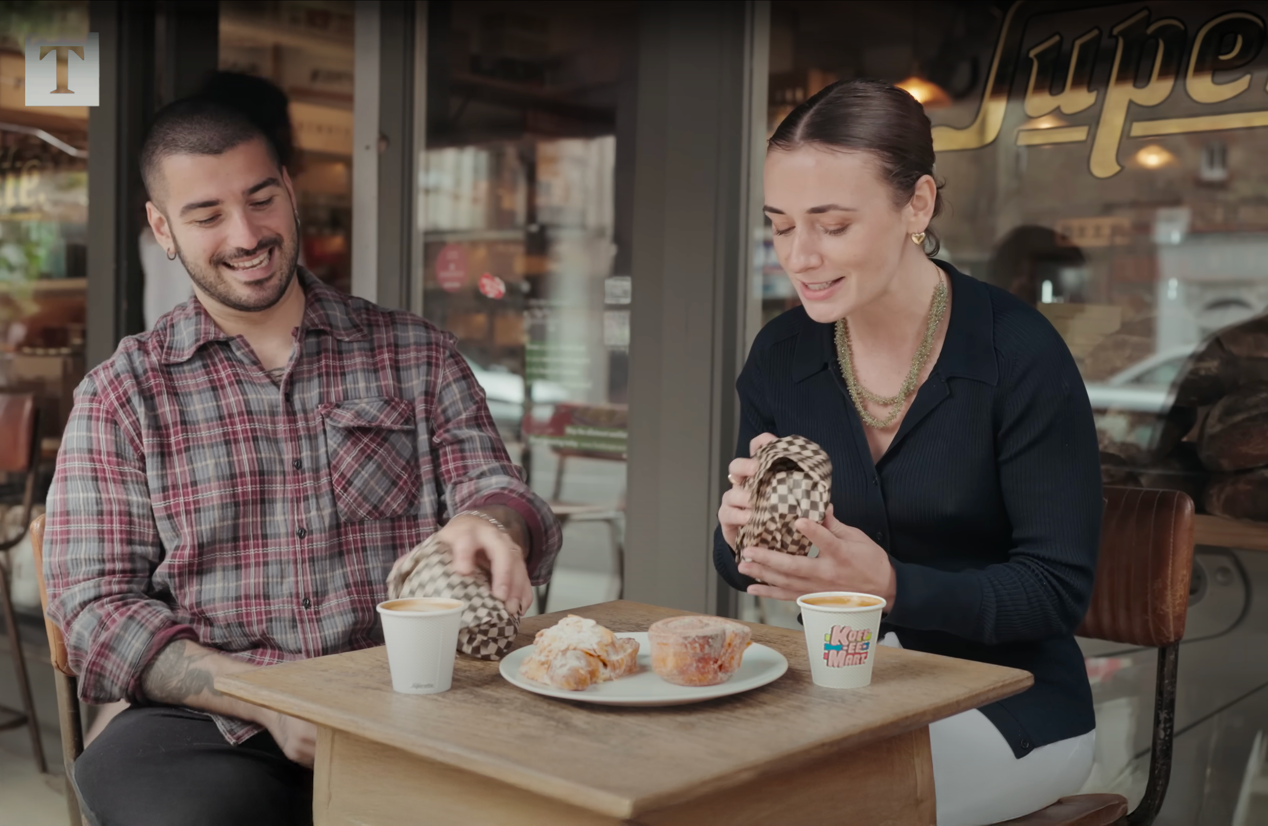 A man and a woman sit at a wooden table outside a cafe, opening checkered paper bags and smiling. There are two cups of coffee and a plate with pastries on the table.