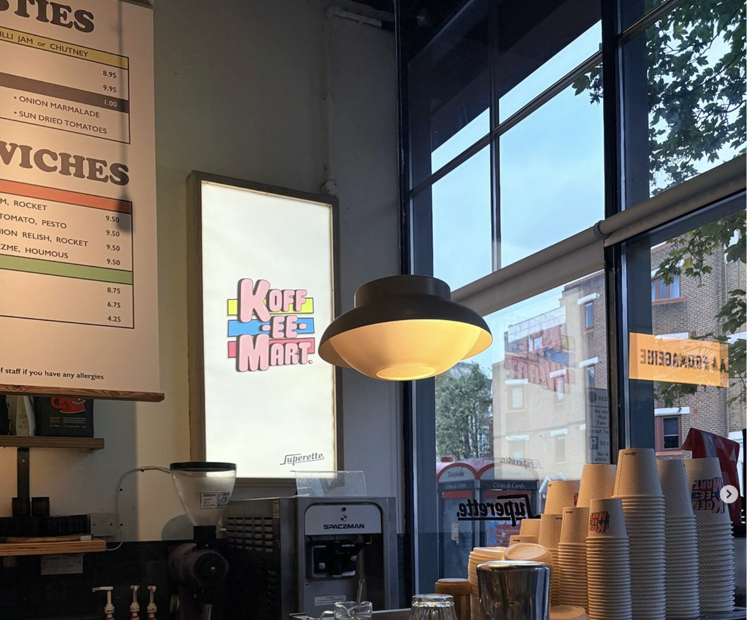 Inside a cafe, with a coffee machine, stacked paper cups, a lighted sign reading 'KOFFEE MART,' a menu on the wall, and large windows showing a street view outside.