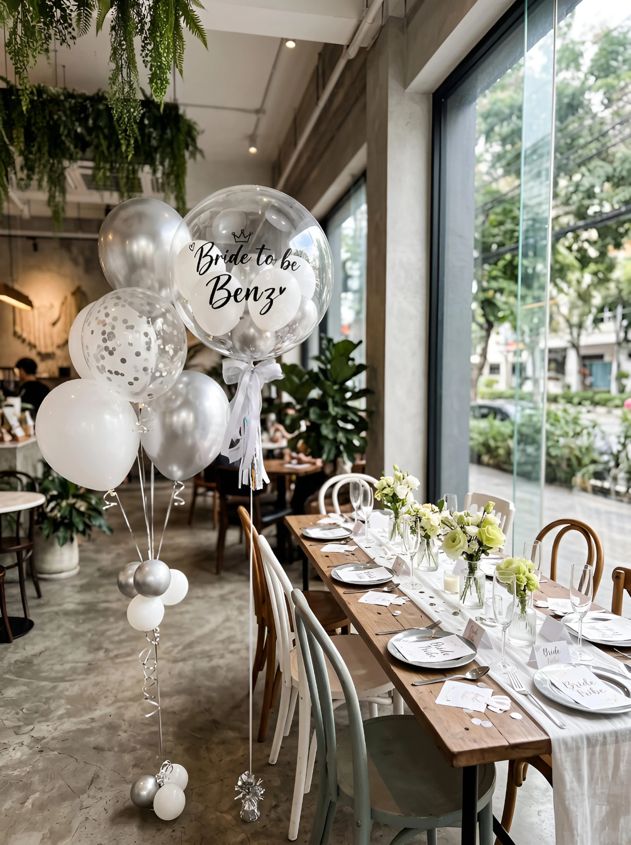 Indoor wedding reception with a long wooden table decorated with white flowers, plates, cutlery, and glasses, and white balloons with one balloon displaying the text 'Bride to be Benz' and a small crown illustration.