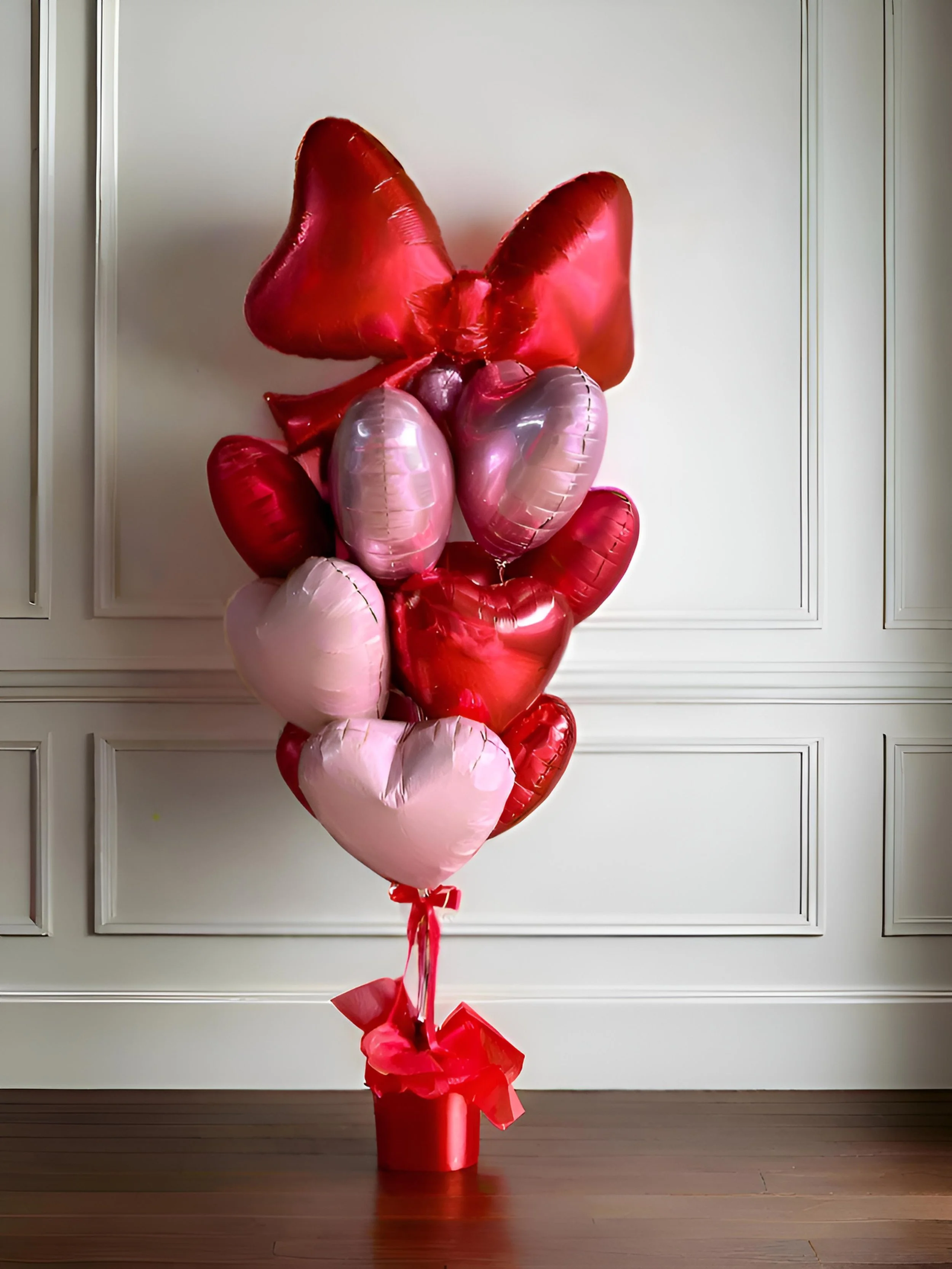 A bouquet of red and pink heart-shaped and butterfly-shaped foil balloons arranged in a cluster, tied to a red foil-wrapped weight, set against a white wall with decorative paneling.