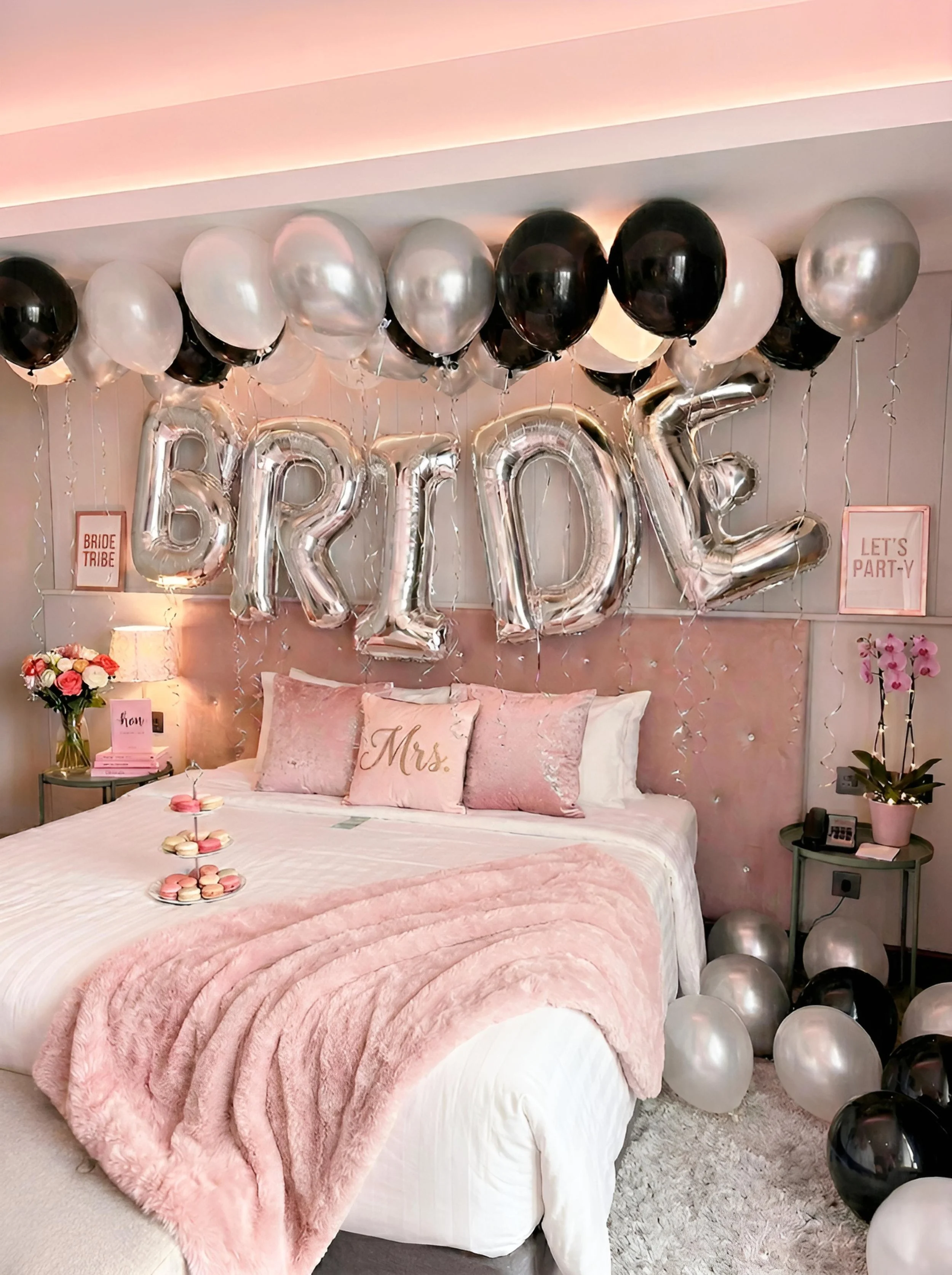 Decorated bedroom with pink and silver balloons spelling "BRIDE" and additional balloons on the floor. The bed has pink pillows, a pink plush blanket, and a tray of pink and white macarons. There are signs reading "BRIDE TRIBE" and "LET'S PARTY!" on the wall, along with flowers, pink framed artwork, and string lights.