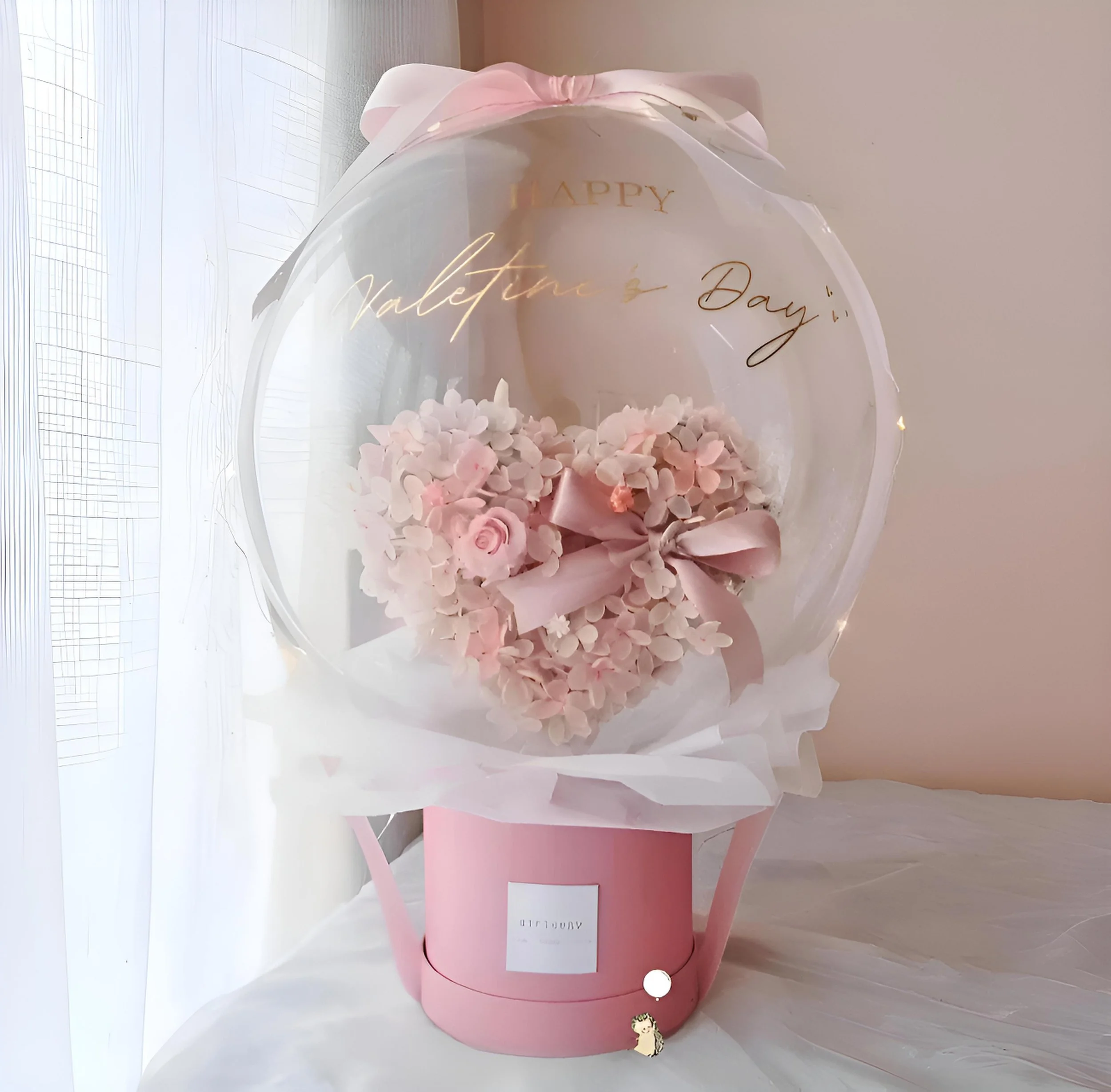 Pink Valentine's Day flower arrangement with roses and hydrangeas inside a clear balloon, attached to a pink gift box.