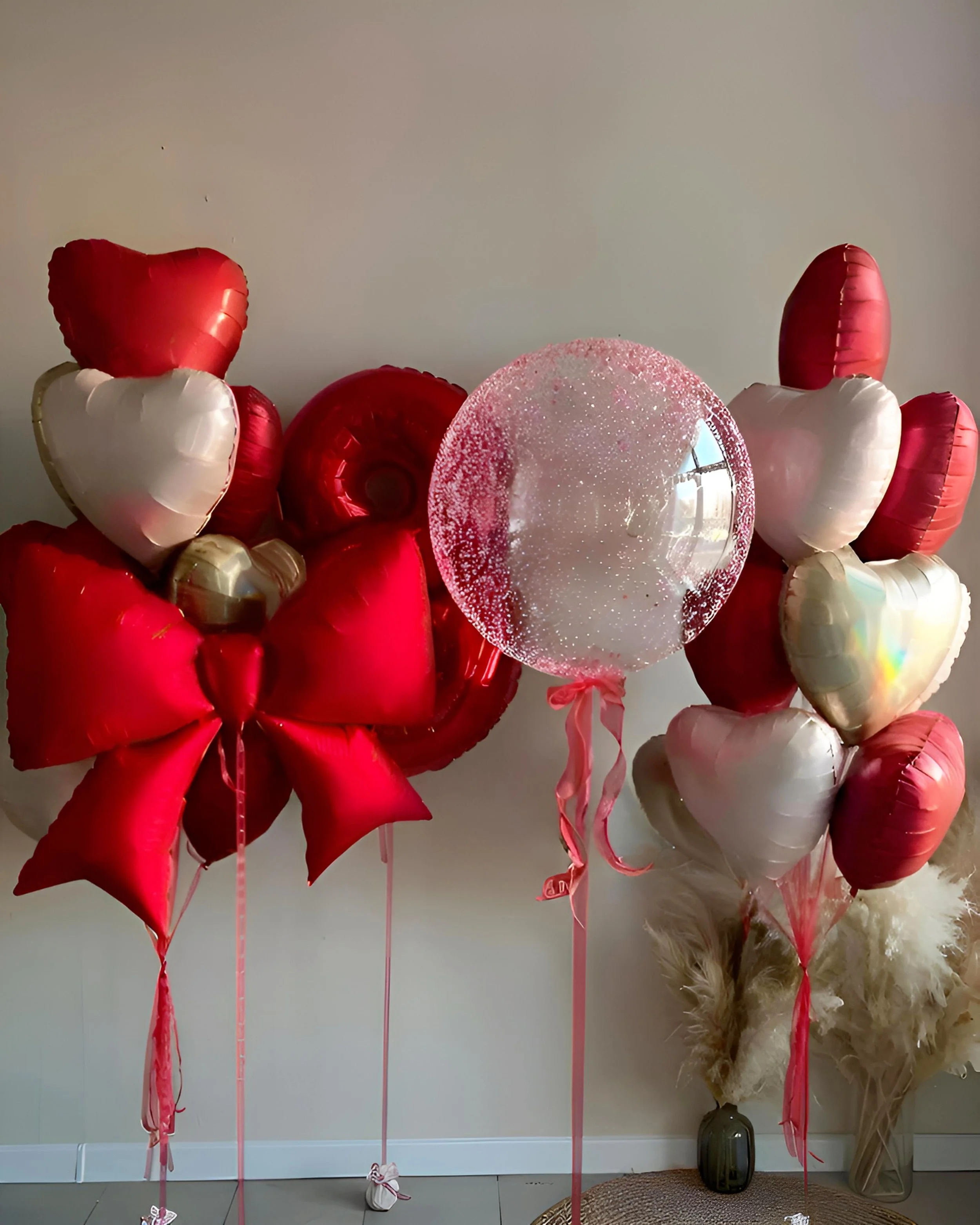 Several balloons in red, white, and pink, including heart-shaped and round ones, arranged with ribbons in a room against a plain wall.
