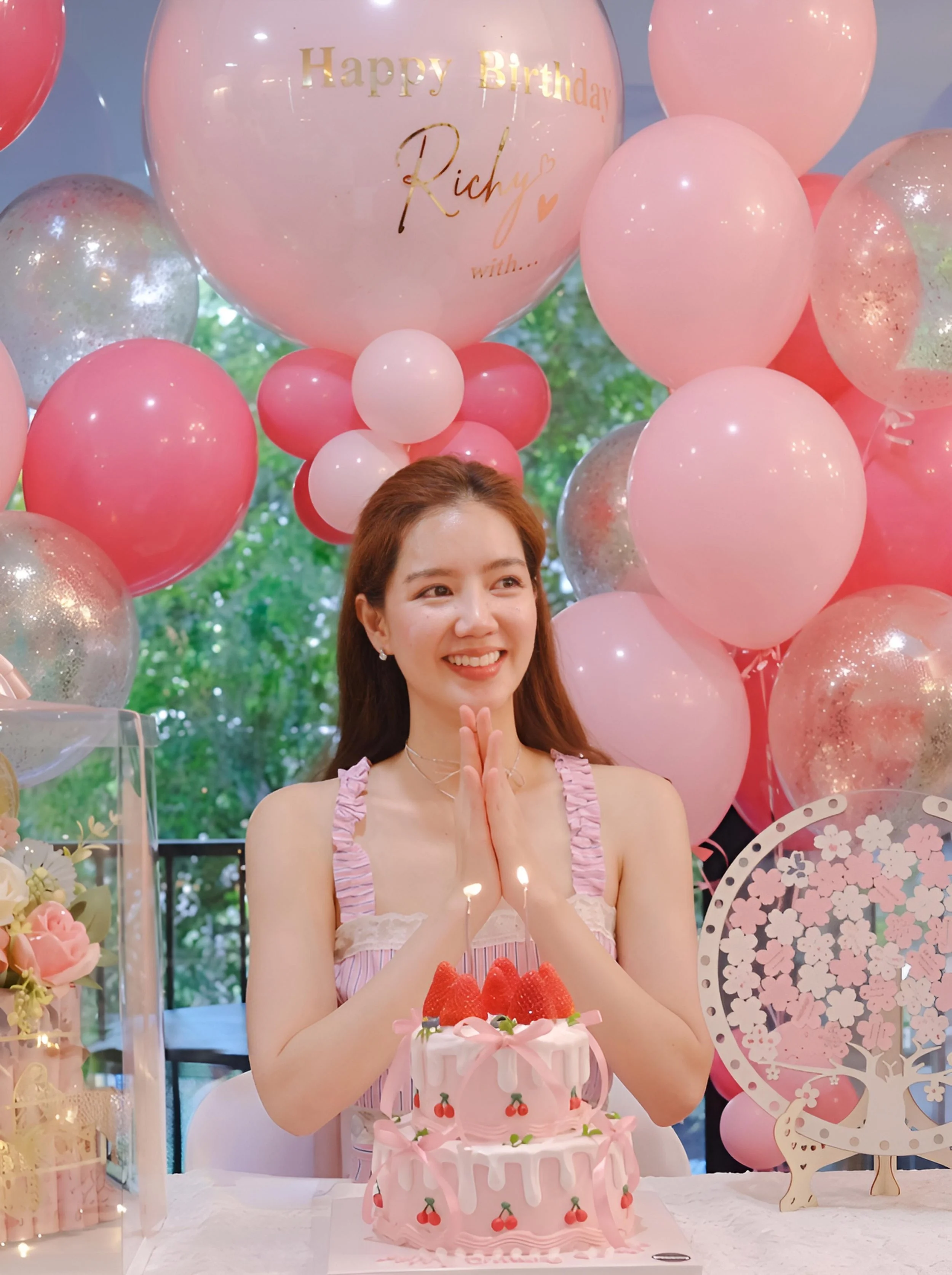 A young woman with red hair smiles in front of a pink birthday cake decorated with strawberries and pink ribbons, surrounded by pink and confetti balloons, with a large pink balloon with gold lettering that says 'Happy Birthday Richy' in the background.