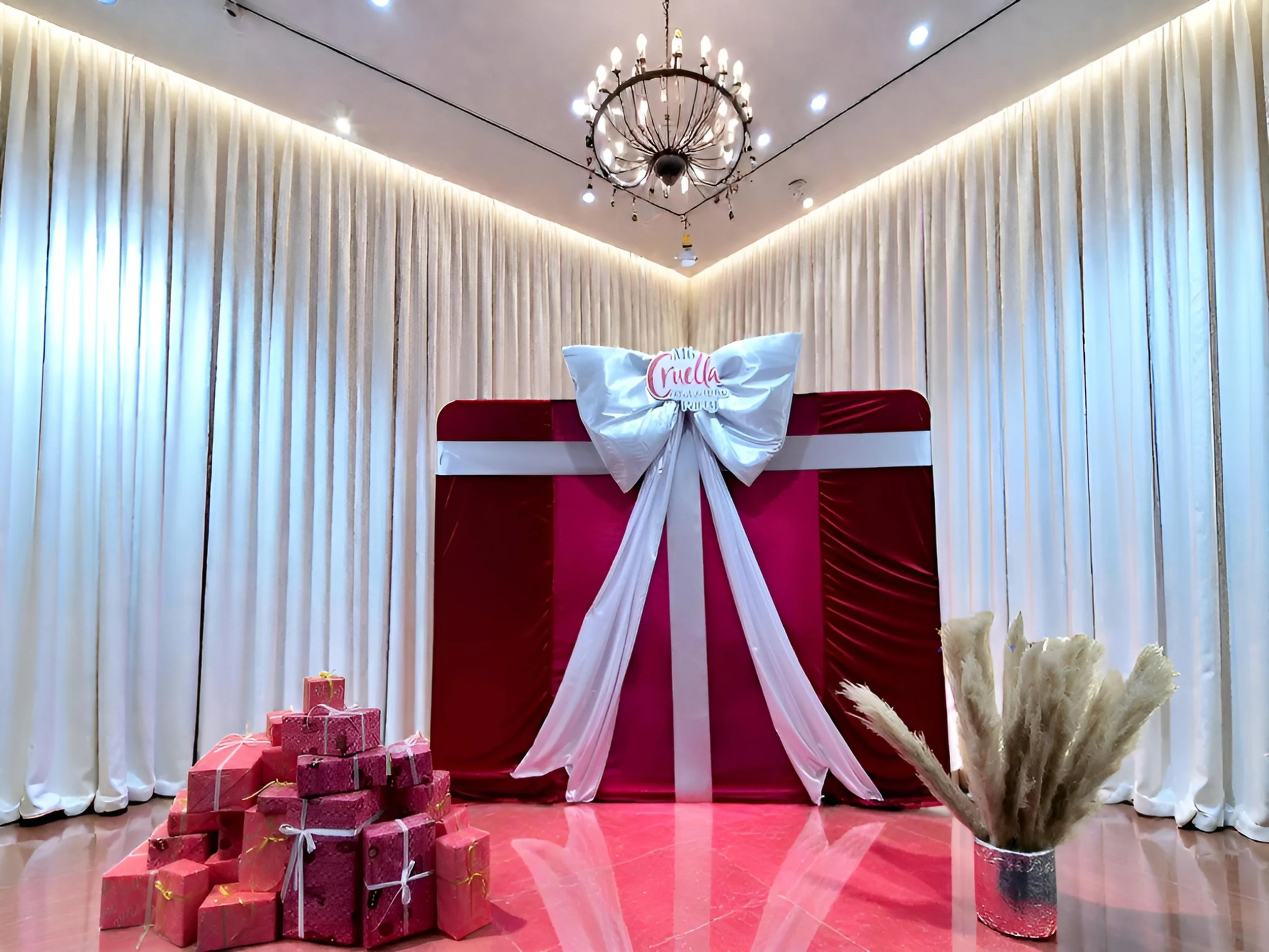 A decorated indoor space with white curtains, a chandelier, and a large pink gift box with a white ribbon and bow. There are decorated pink gift boxes stacked on the left and a bunch of pampas grass in a silver vase on the right.